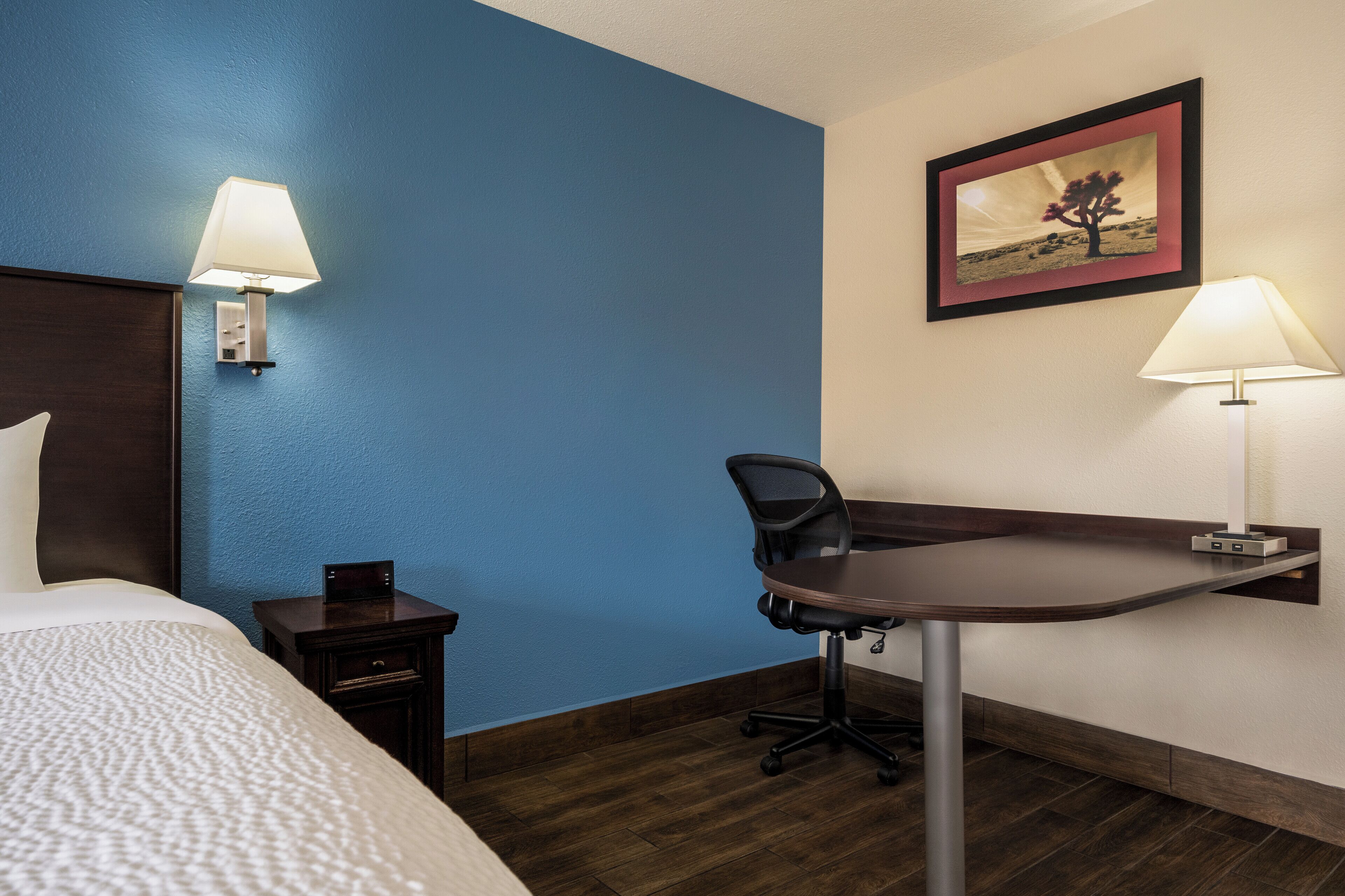 A hotel room with a bed, a desk, a chair, a nightstand, and two wall-mounted lamps.