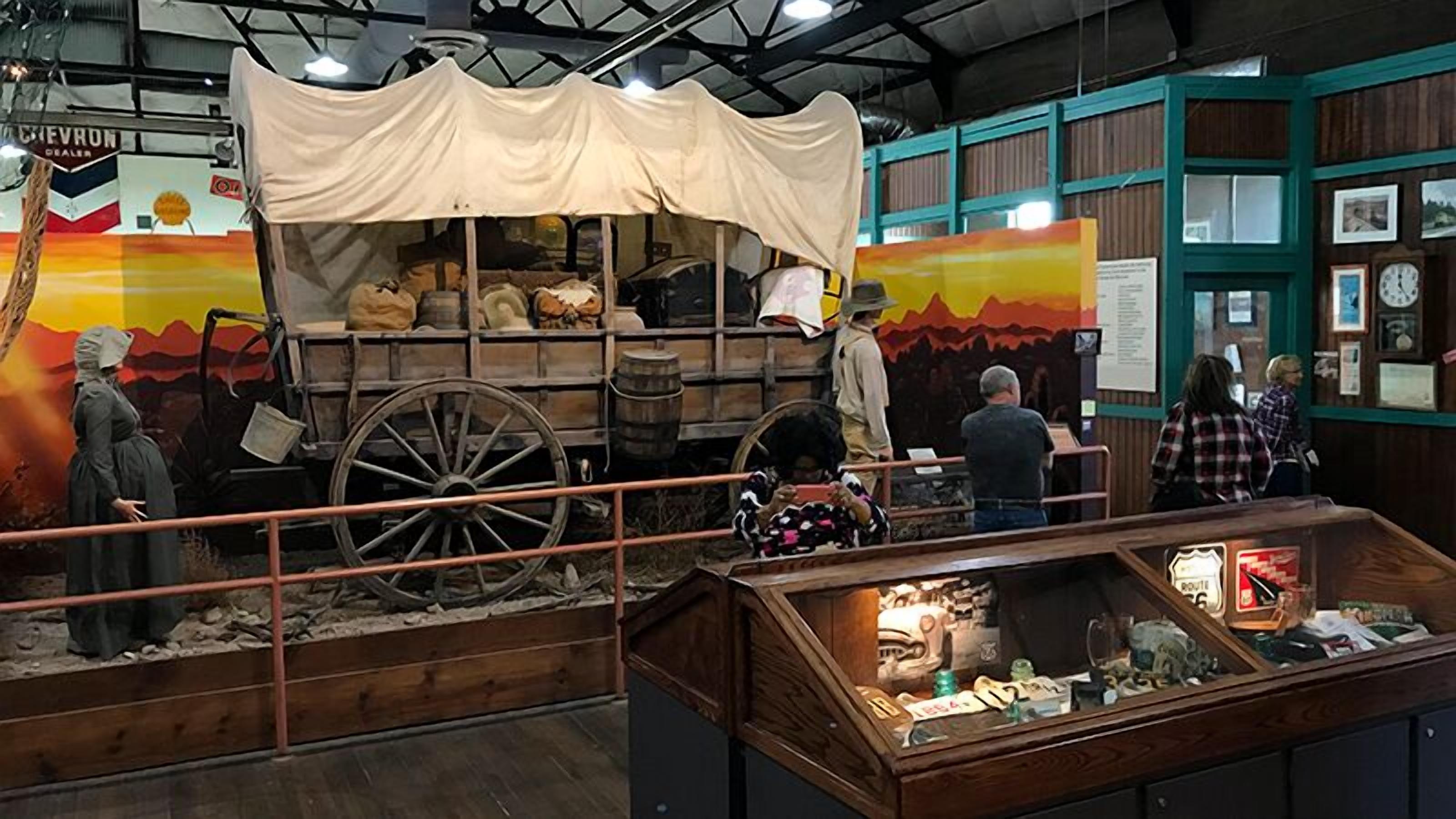 Una exposición histórica con un carro cubierto, barriles de madera y una vitrina con diversos objetos.