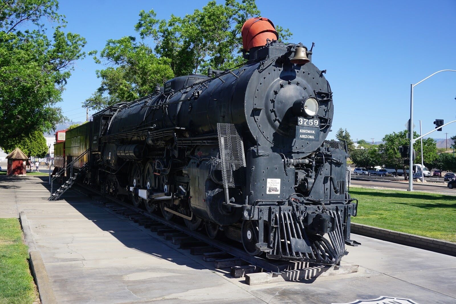 Una locomotora de vapor grande e inmóvil con el número 3759, situada sobre una vía de tren en un espacio público con árboles y un cielo despejado al fondo.