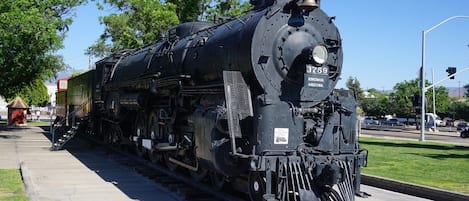Una locomotora de vapor grande e inmóvil con el número 3759, situada sobre una vía de tren en un espacio público con árboles y un cielo despejado al fondo.