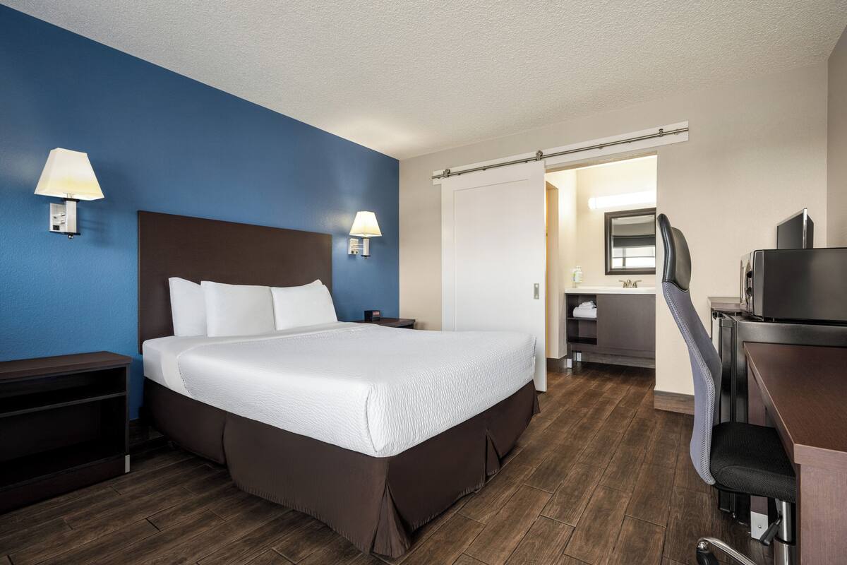 A hotel room with a large bed, a desk, and a chair. There are two lamps on the wall, a mirror, and a door leading to a bathroom.