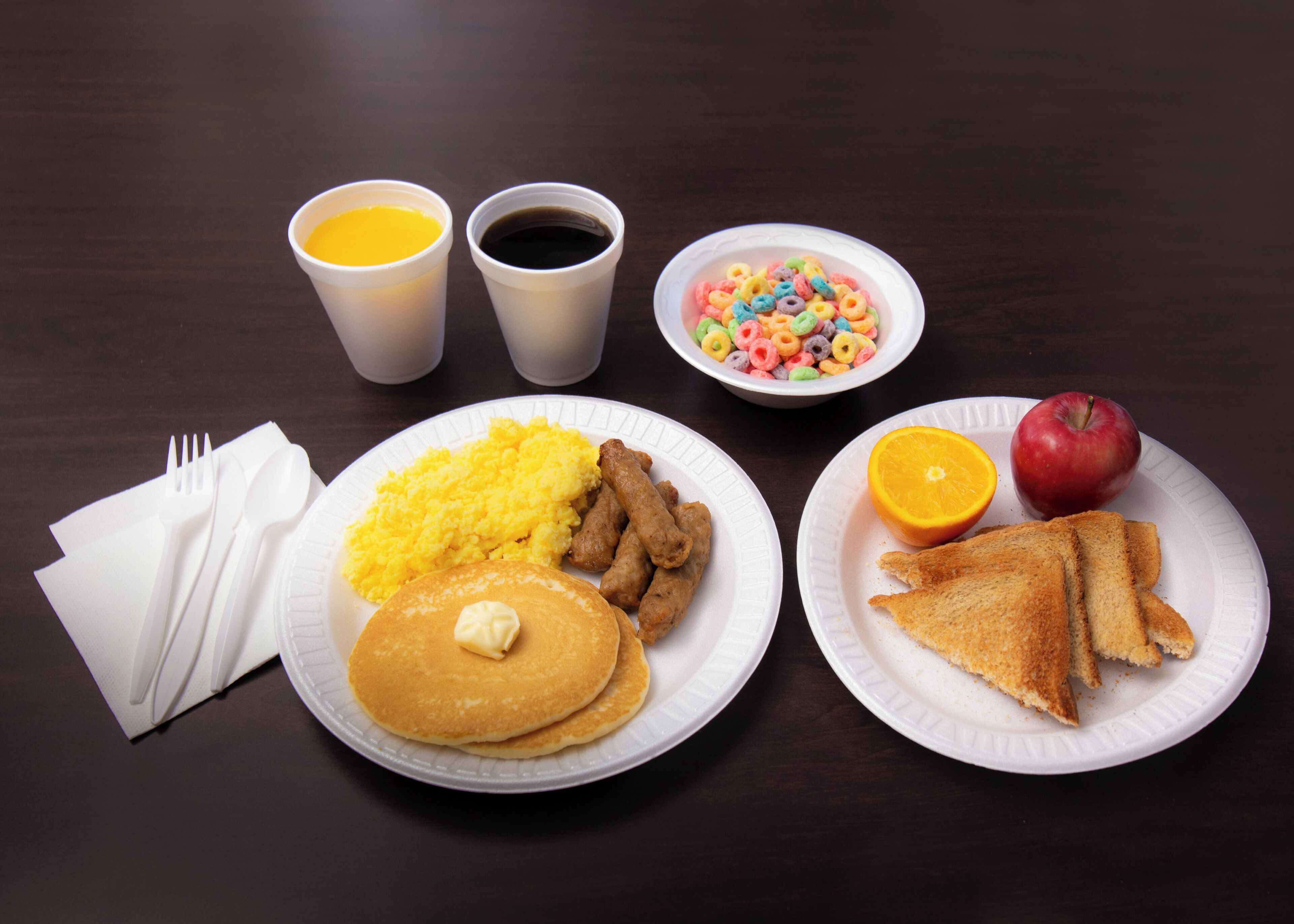 Ein Teller mit Pfannkuchen, Rührei, Würstchen und einer Schüssel Müsli.