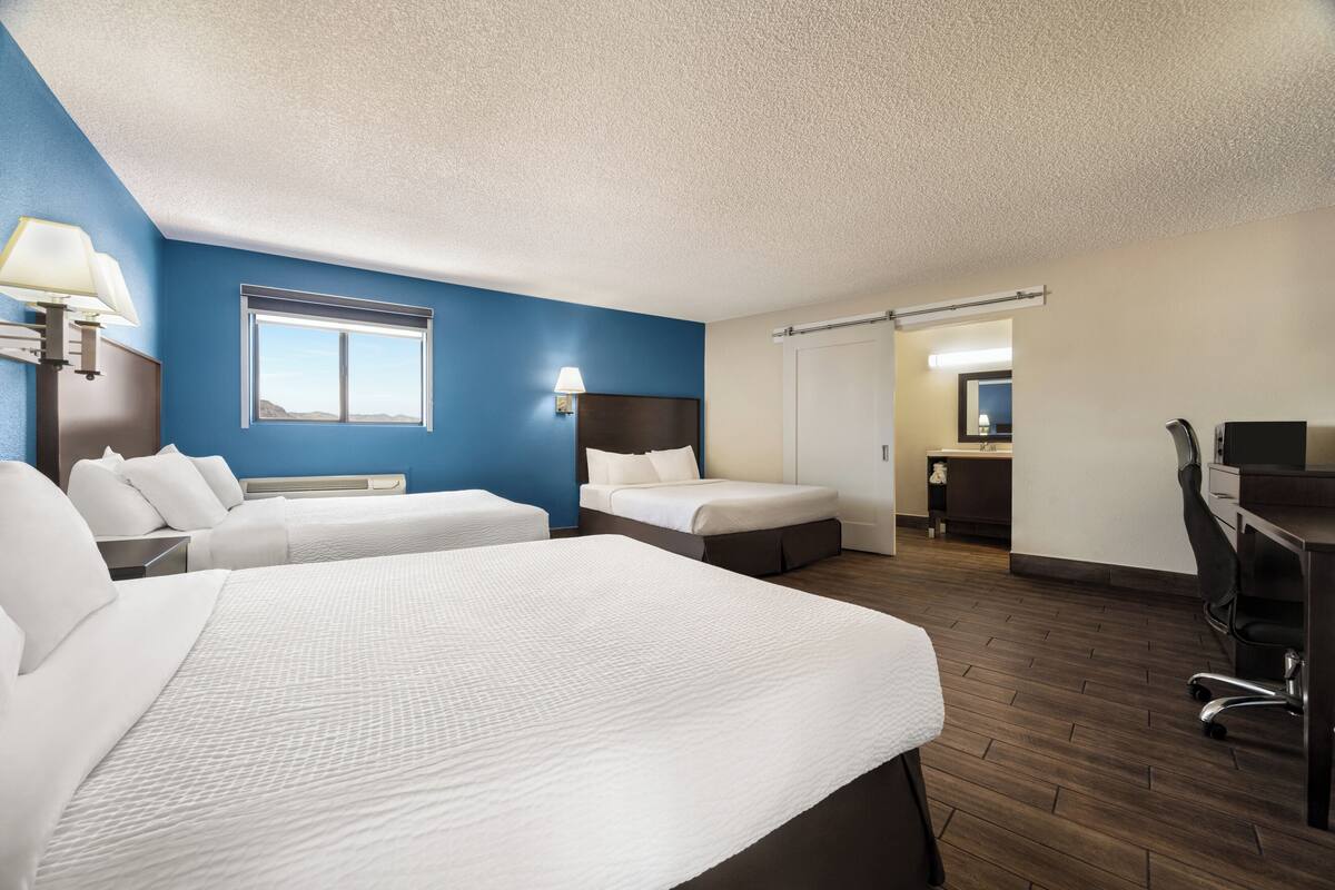 A hotel room with two beds, a desk, a chair, a window, and a bathroom visible through an open door.
