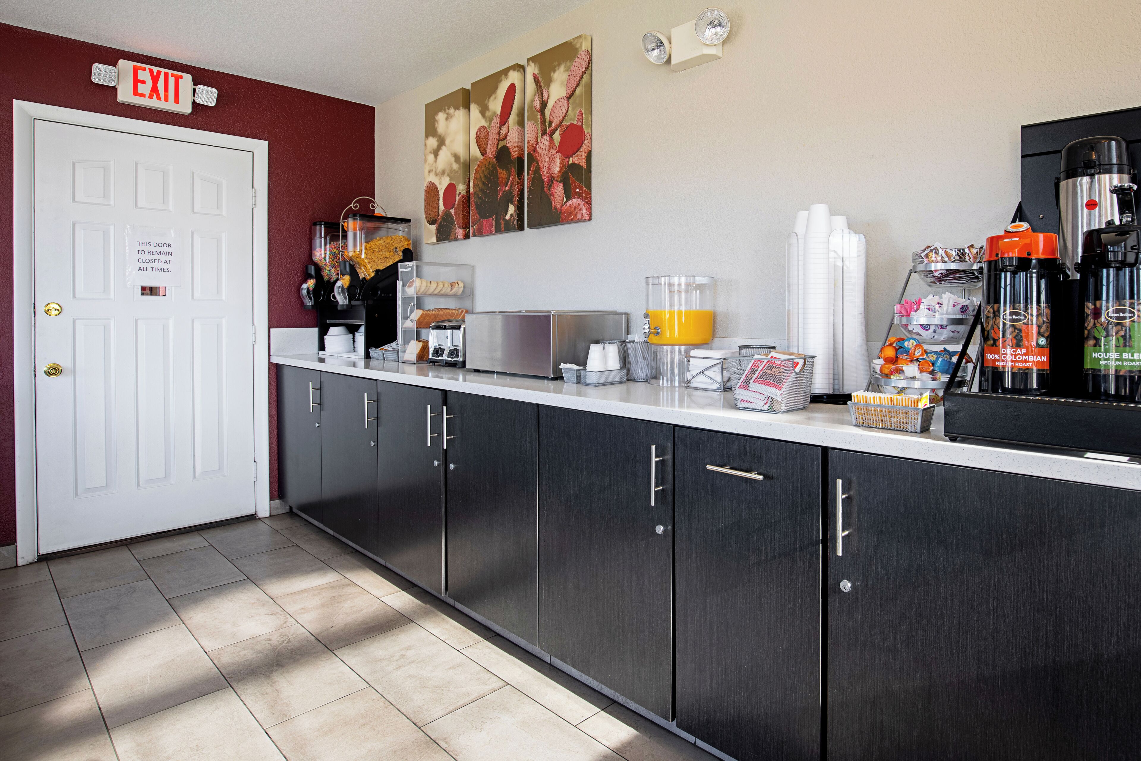 A clean, modern reception area with a white door marked 'EXIT', a coffee machine, a fruit bowl, and a coffee station.