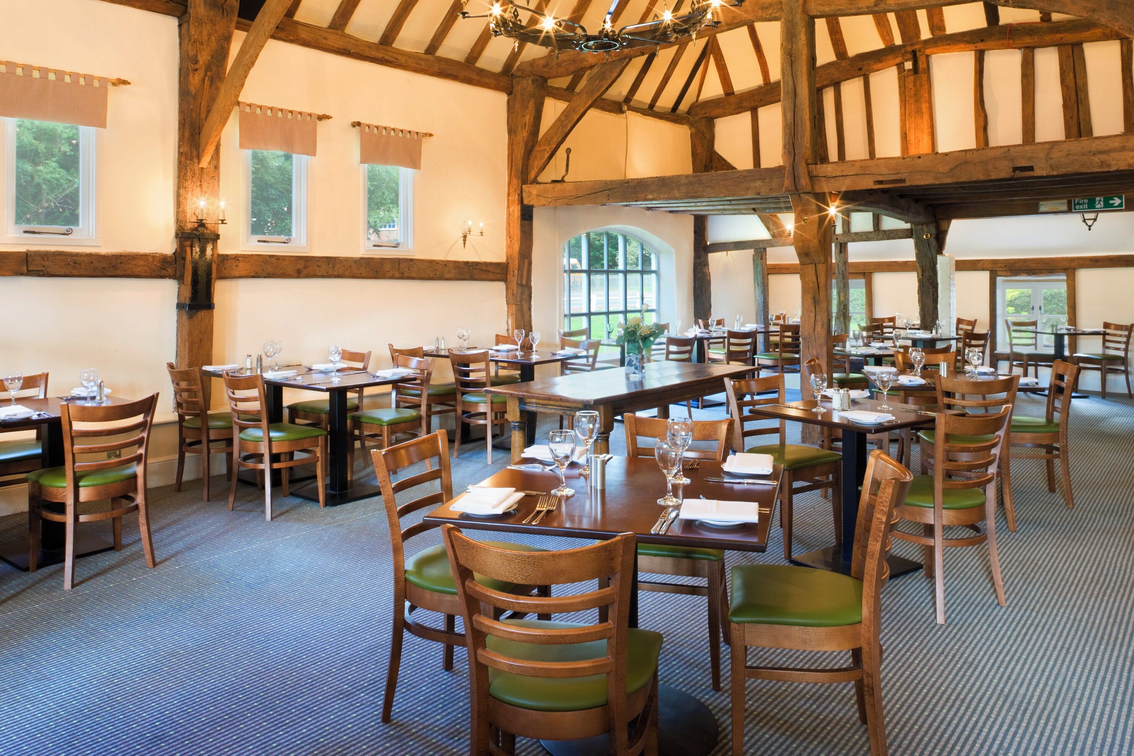 A large dining hall with multiple tables and chairs, wooden beams on the ceiling, and large windows.