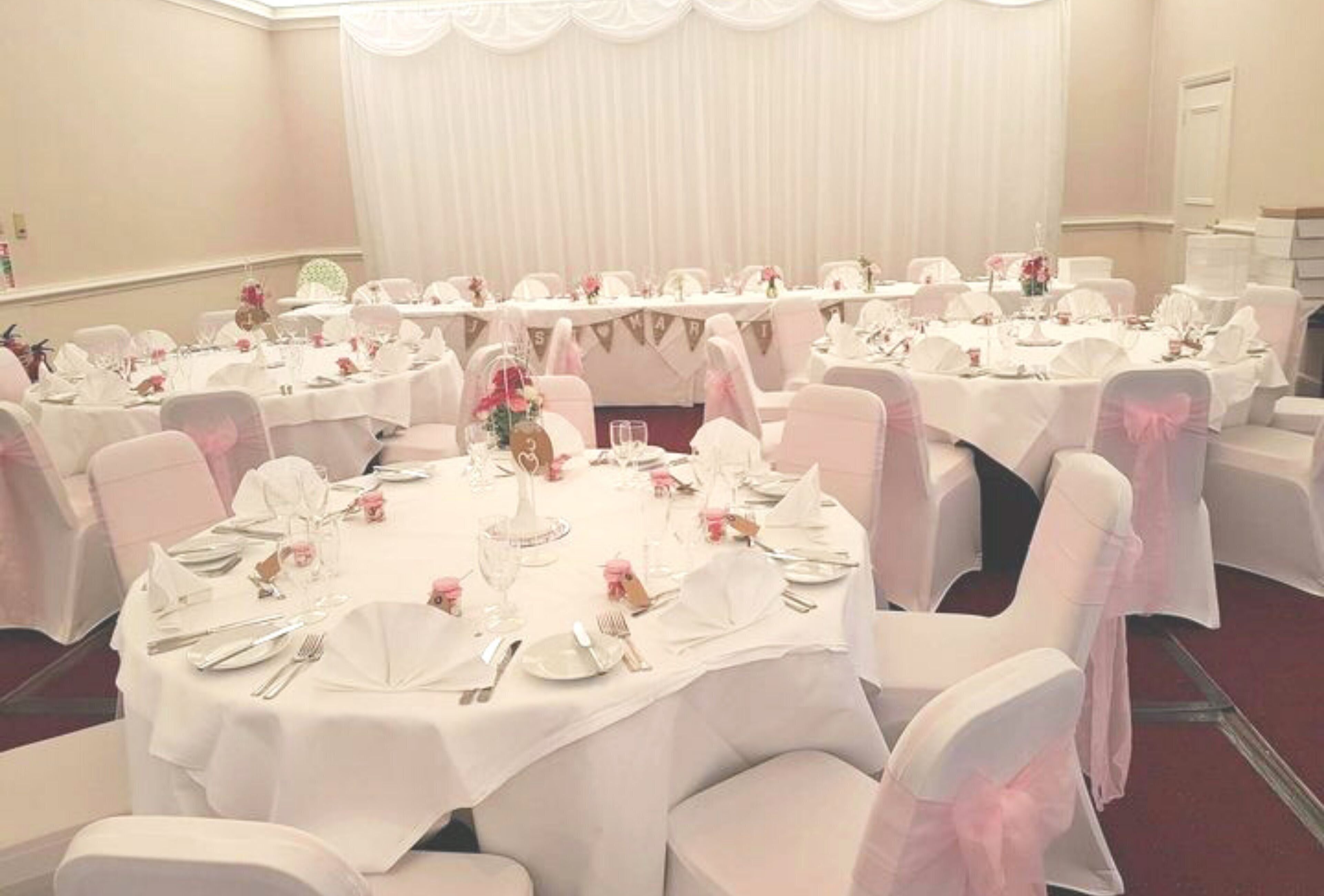 Una sala da banchetto con tavoli rotondi apparecchiati per la cena, tovaglie bianche e sedie con nastri rosa.