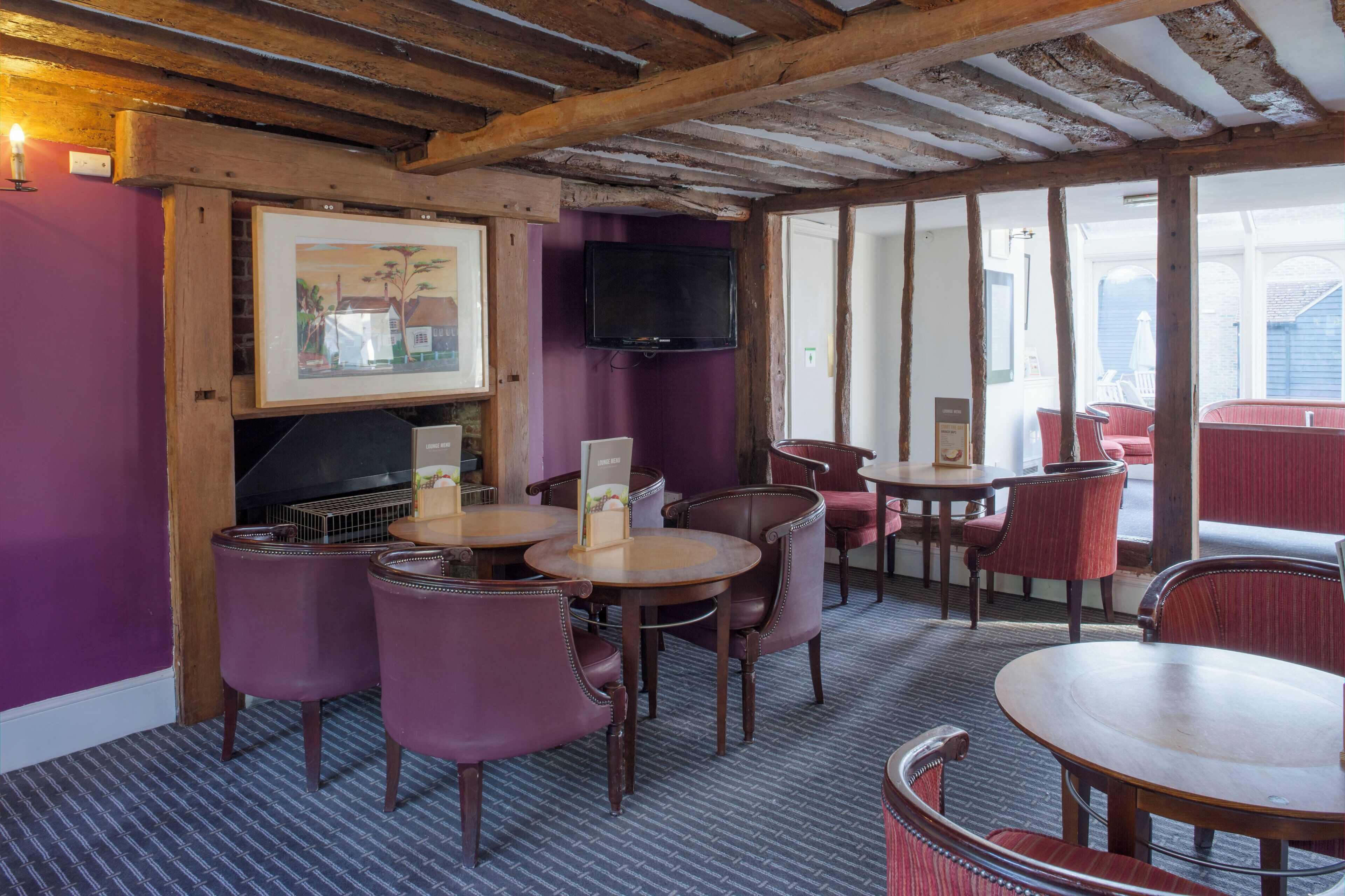 Un restaurant avec des poutres en bois, des murs violets et un téléviseur fixé au mur. On y trouve des tables et des chaises pour les repas.