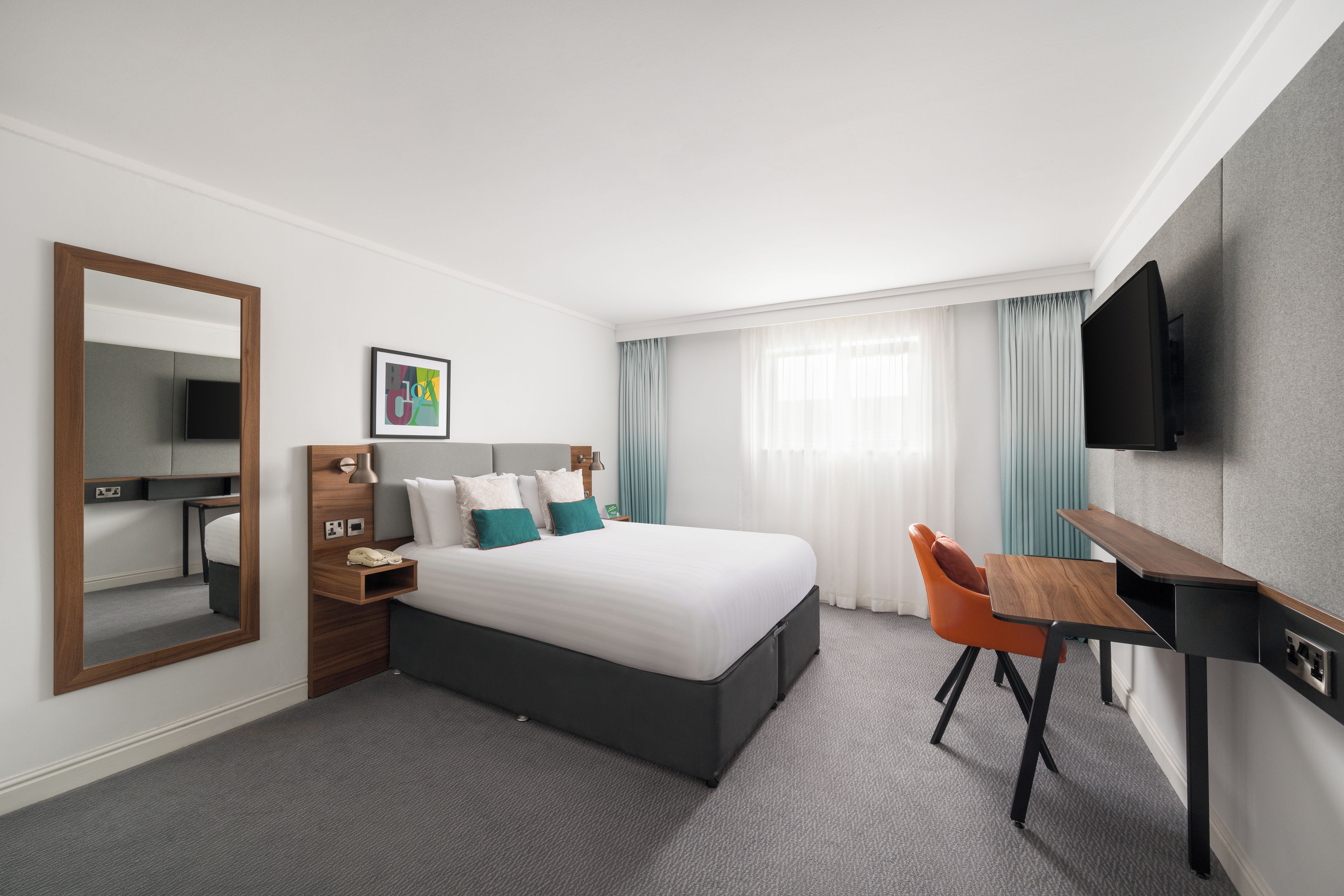 Una habitación de hotel con cama, escritorio, silla, televisor, espejo y ventana con cortinas.
