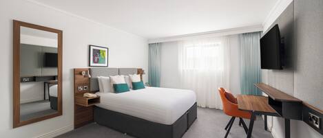 Una habitación de hotel con cama, escritorio, silla, televisor, espejo y ventana con cortinas.