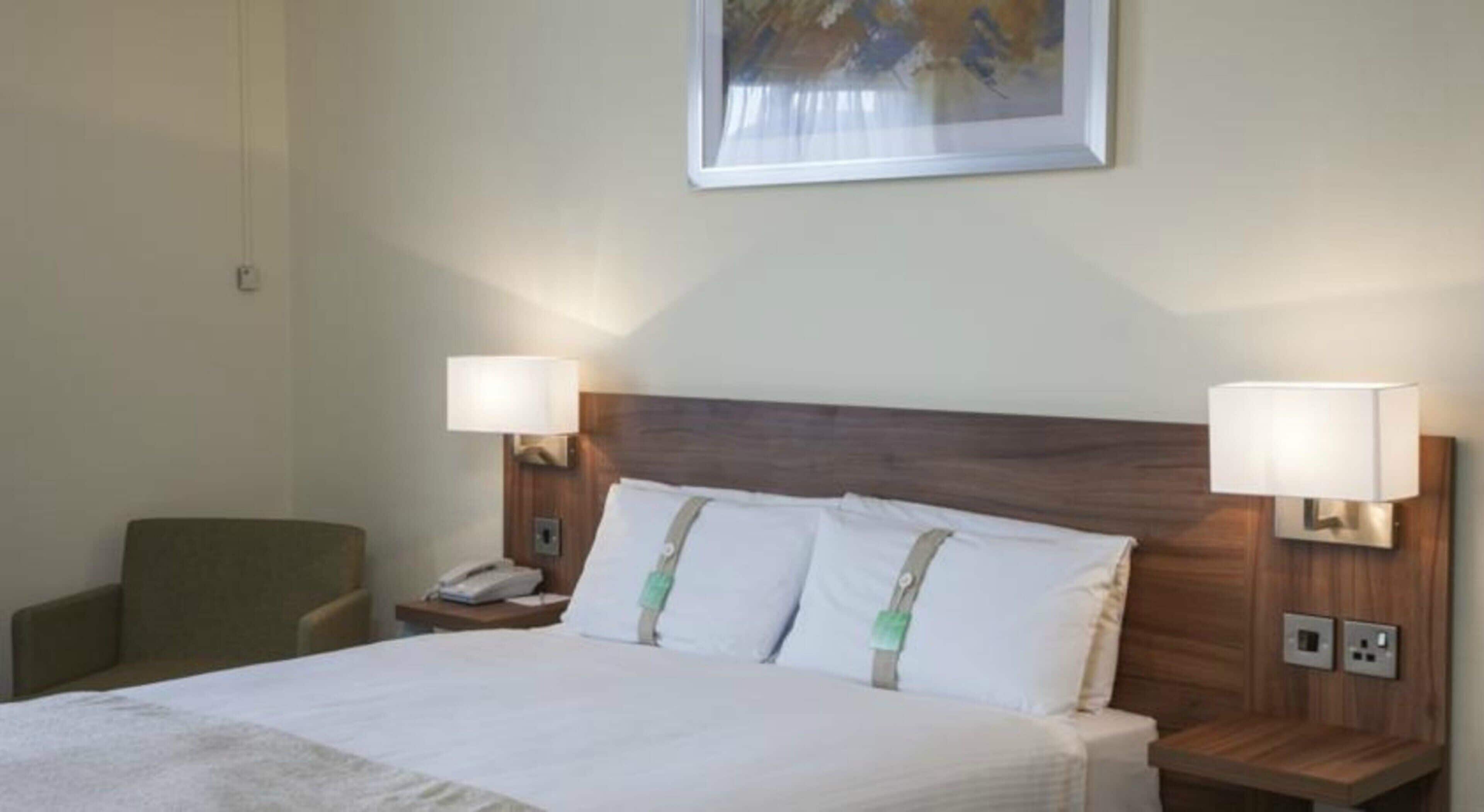 Habitación de hotel con una cama, dos lámparas de noche, una silla, una mesita de noche y un cuadro en la pared.