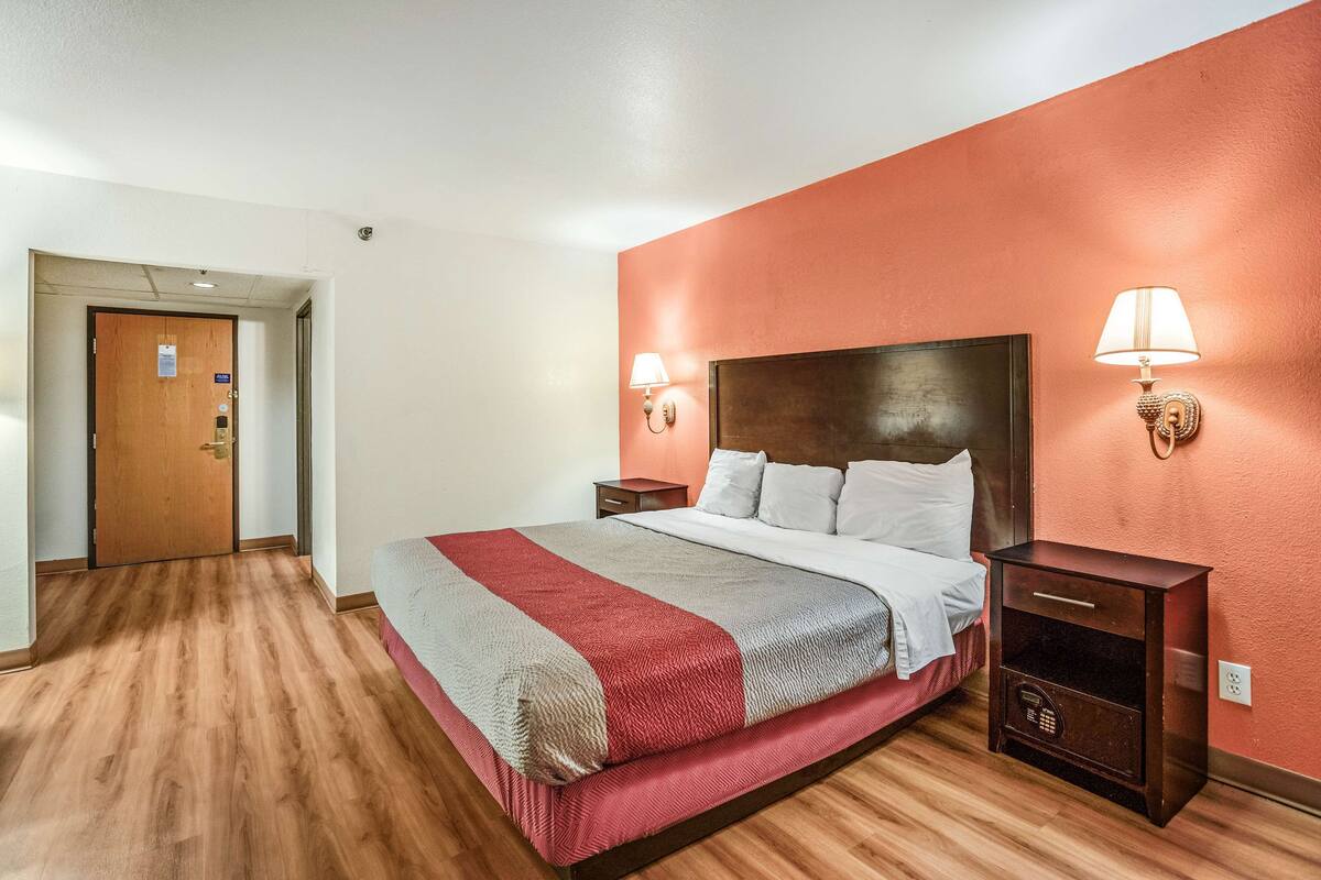 Ein Hotelzimmer mit Holzboden, einem Bett mit roter und grauer Decke, einem Holz-Kopfteil, zwei Nachttischen mit Lampen sowie einer Tür mit Schloss.