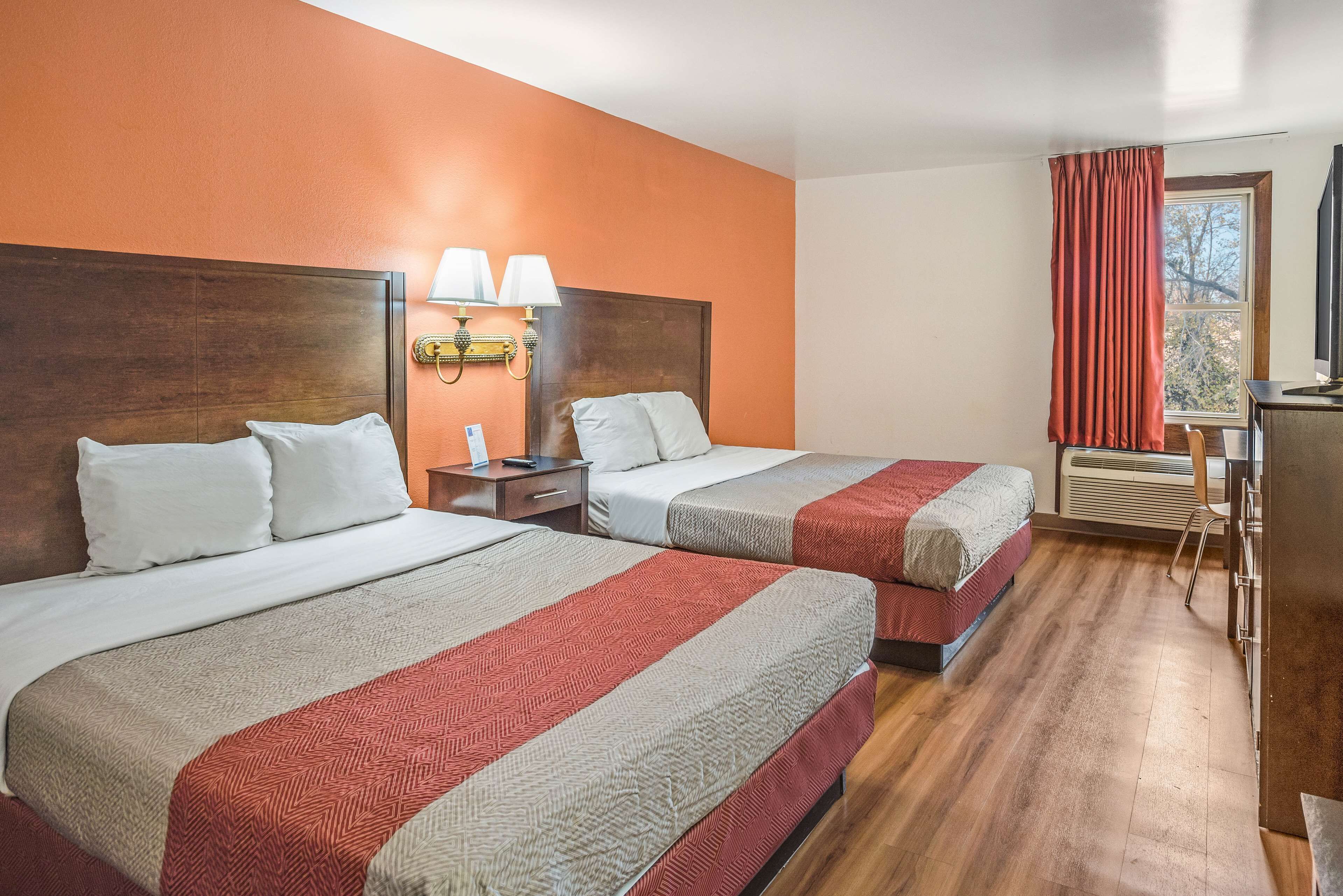 A hotel room with two beds, a nightstand, a lamp, a window with curtains, and a desk with a chair.