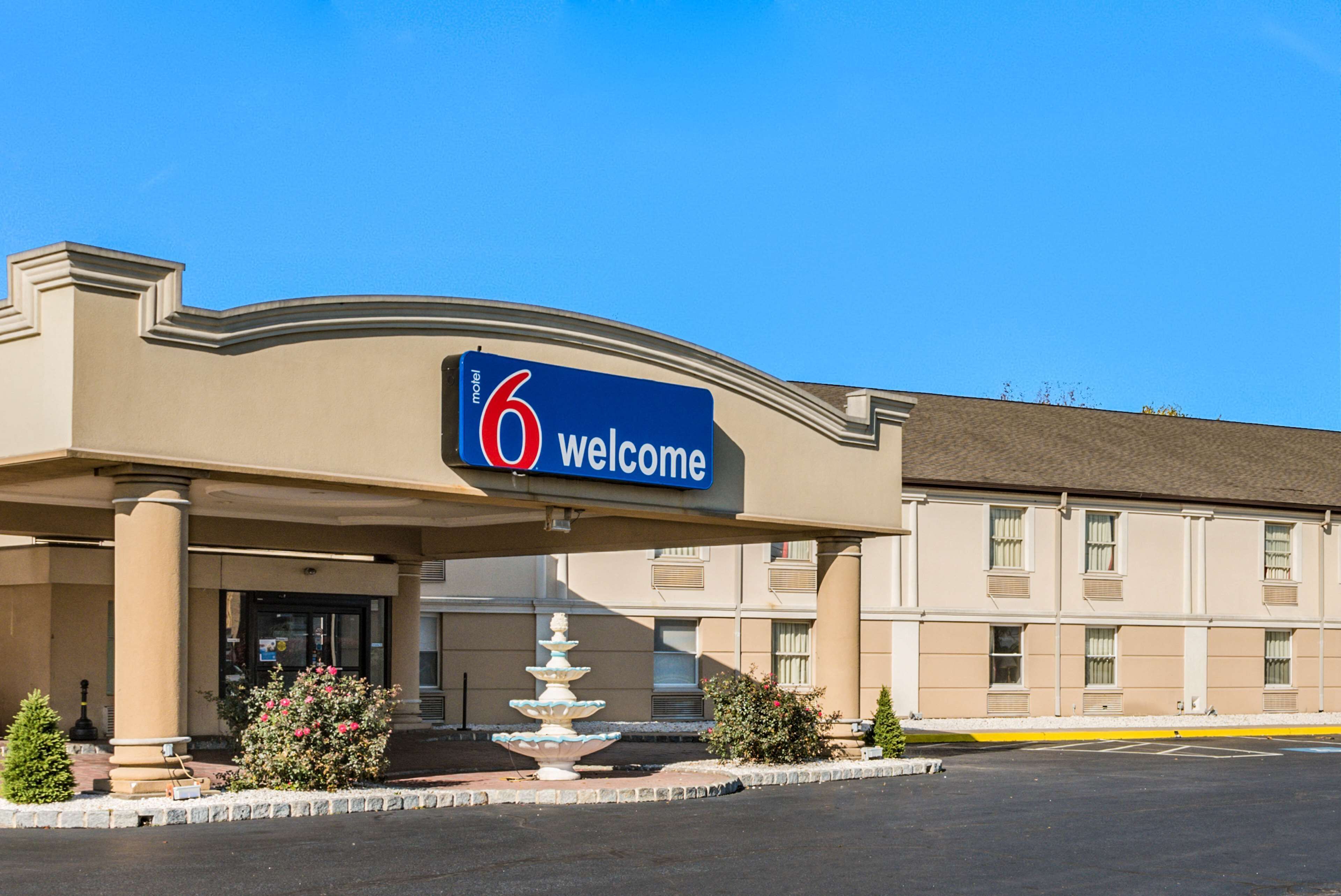 Entrada de hotel con un letrero luminoso que dice '6 Welcome', una fuente y una zona de estacionamiento.