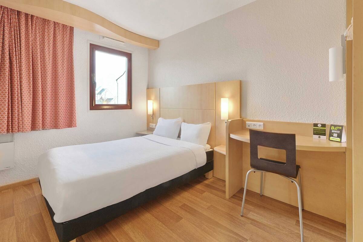A hotel room with a single bed, a desk with a chair, a window with curtains, and a wall-mounted lamp.