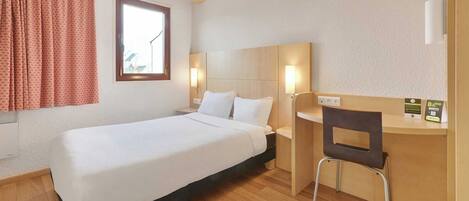A hotel room with a single bed, a desk with a chair, a window with curtains, and a wall-mounted lamp.