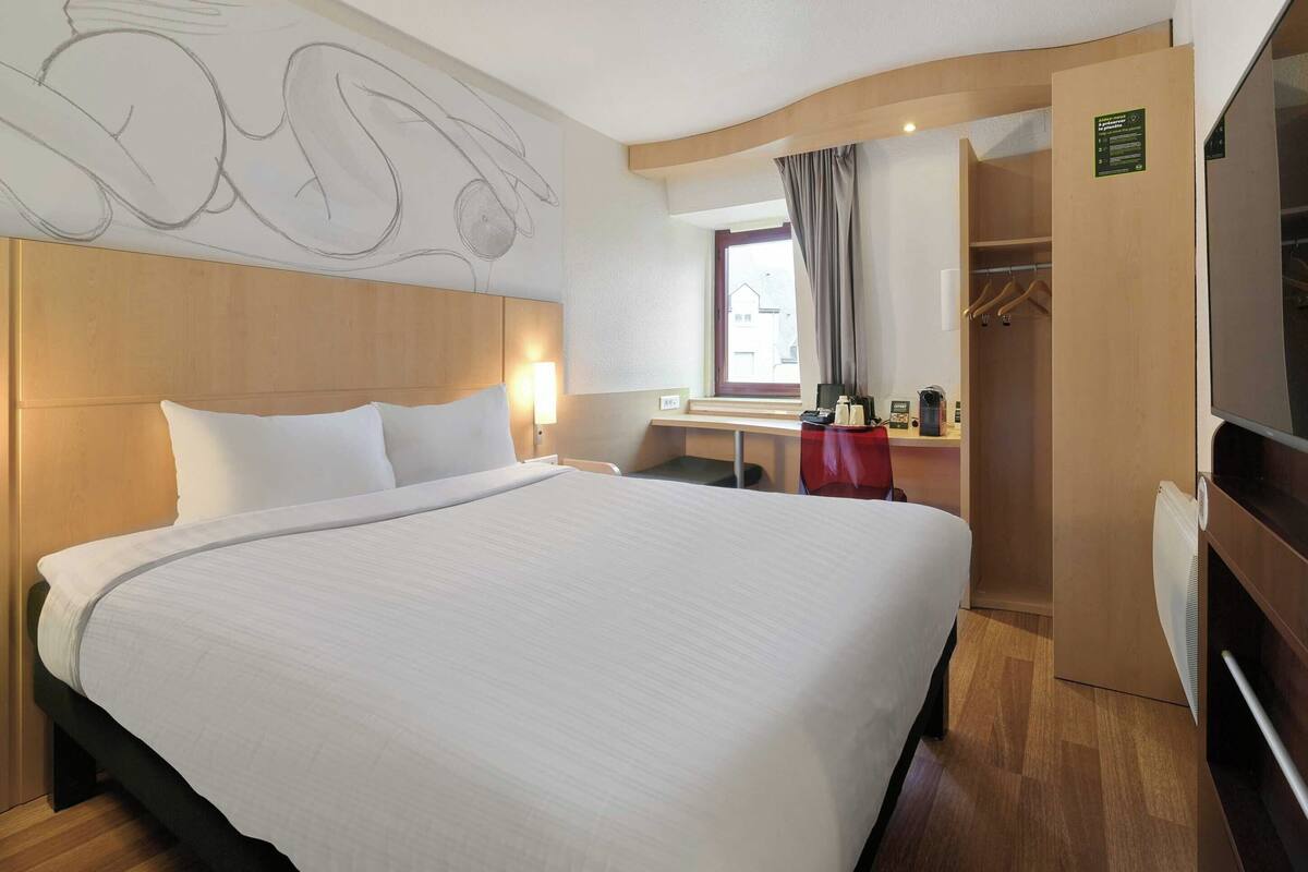 A hotel room with a large bed, a wooden headboard, a desk with a chair, a TV, and a wardrobe.