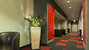 Un couloir d’hôtel avec un tapis à carreaux, une plante en pot, une porte rouge et une poubelle noire.