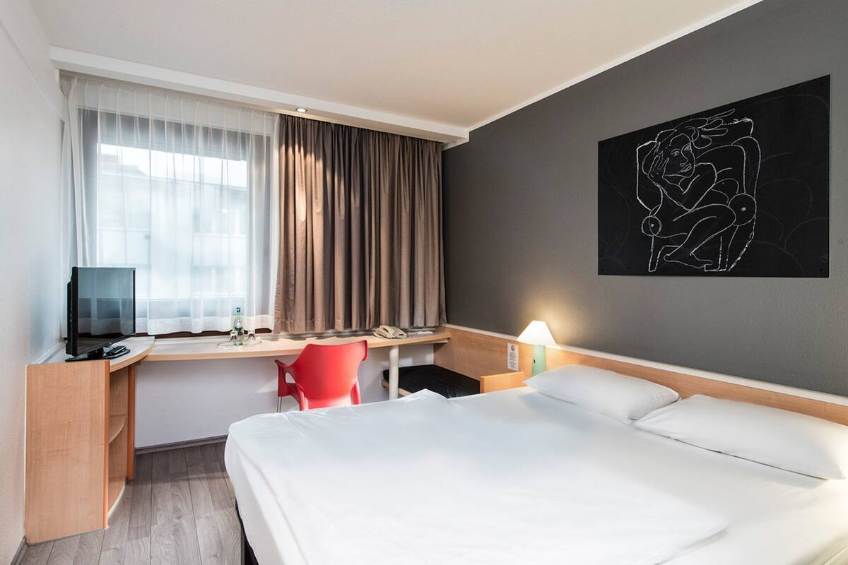 A hotel room with a bed, a desk with a computer, a chair, a window with curtains, and a wall art piece.