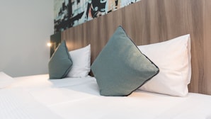 Un lit bien fait, avec du linge de lit blanc et deux coussins turquoise, adossé à une tête de lit en bois sur laquelle est peint un décor représentant un paysage urbain.