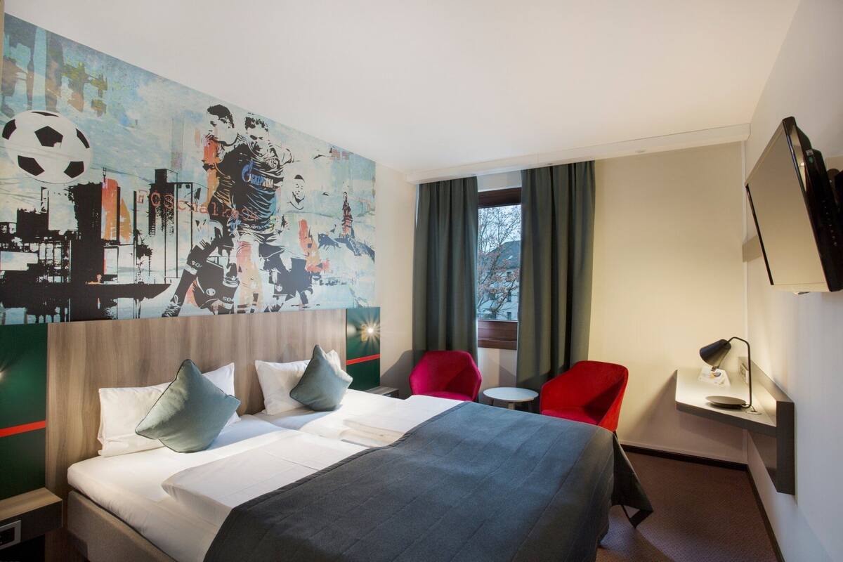 A hotel room with a large bed, a red chair, a small table, and a wall-mounted TV.
