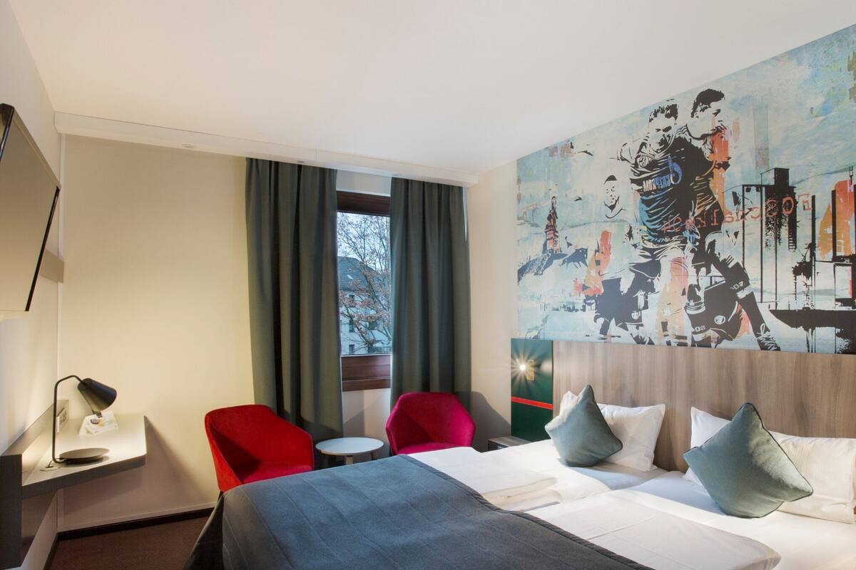 A hotel room with a bed, two red chairs, a small table, a desk with a lamp, and a large painting on the wall.