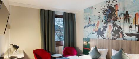 A hotel room with a bed, two red chairs, a small table, a desk with a lamp, and a large painting on the wall.