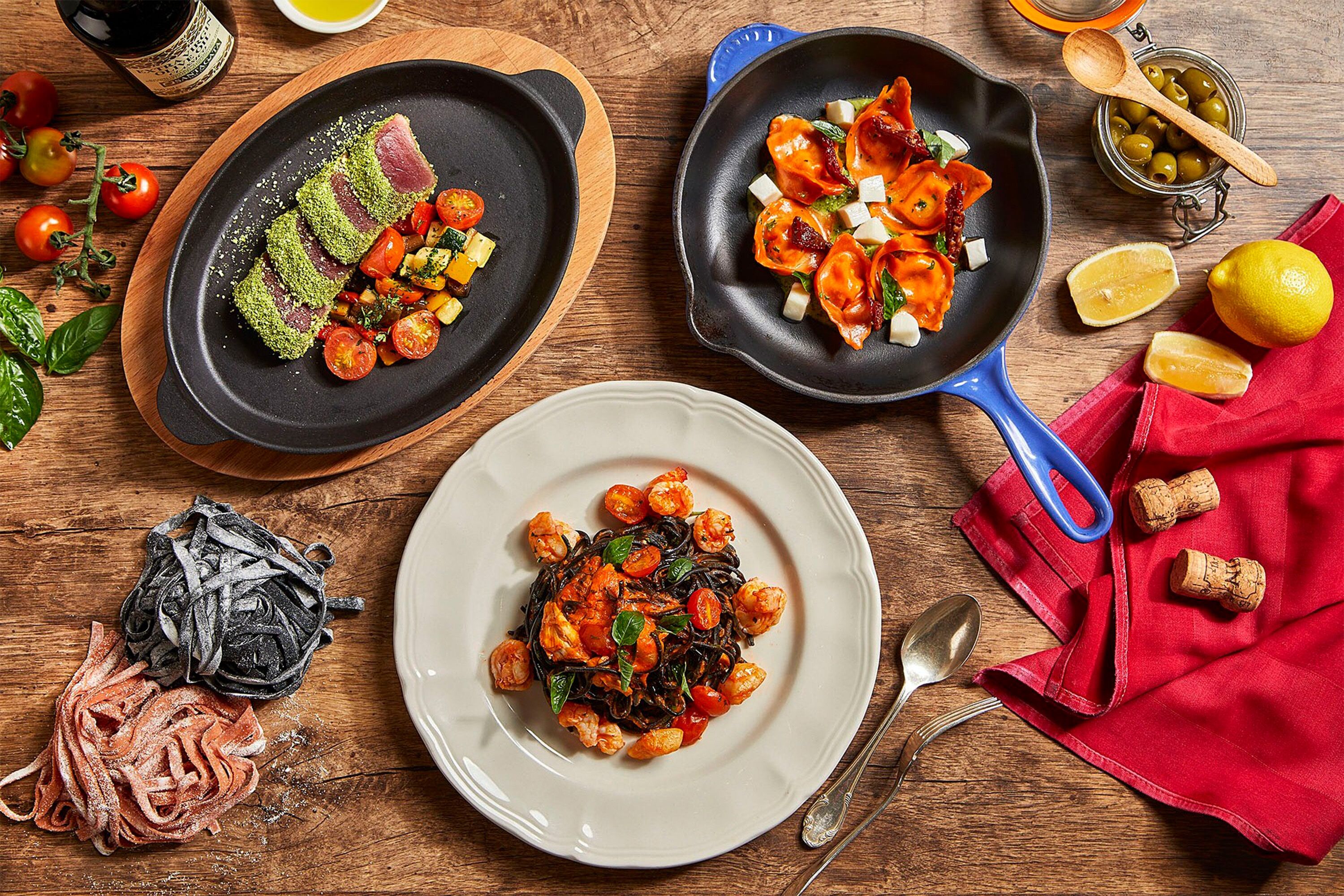 Una mesa de madera con tres platos de comida, una botella de aceite de oliva y un paño rojo.