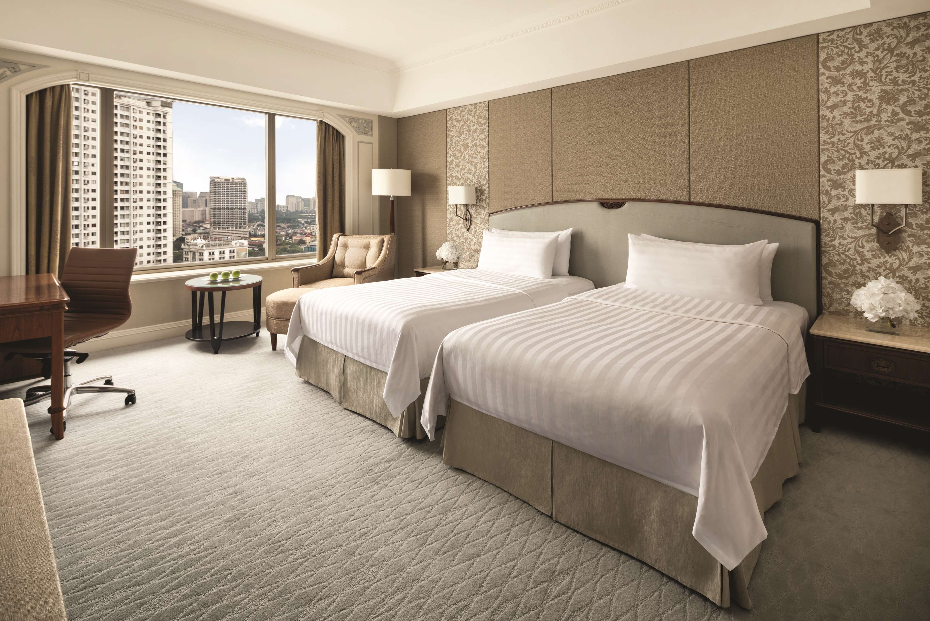 A hotel room with two beds, a desk, a chair, a small table, and a large window offering a city view.