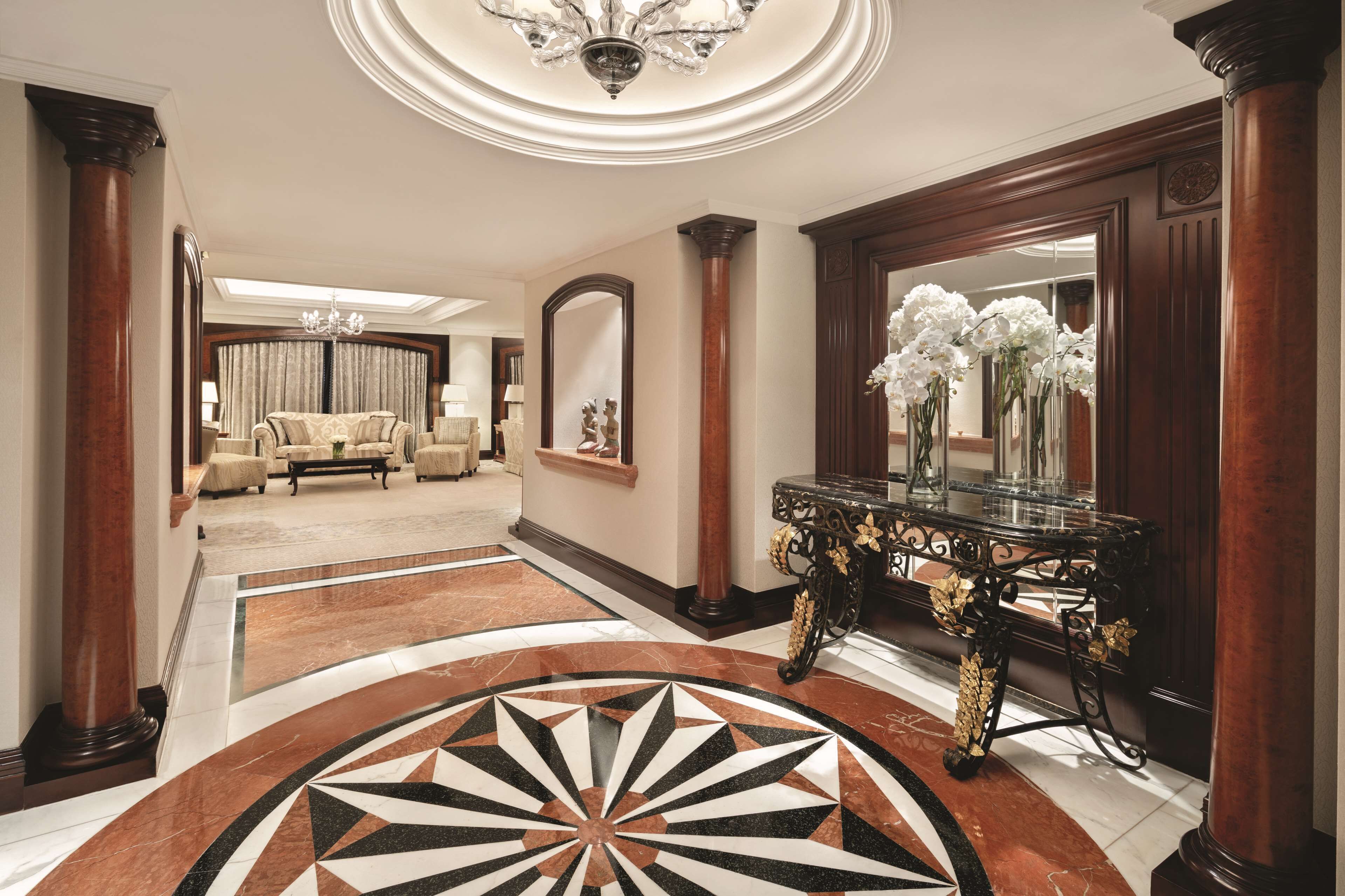 A grand lobby with a large, ornate mirror, a black console table with floral decorations, and a patterned floor.