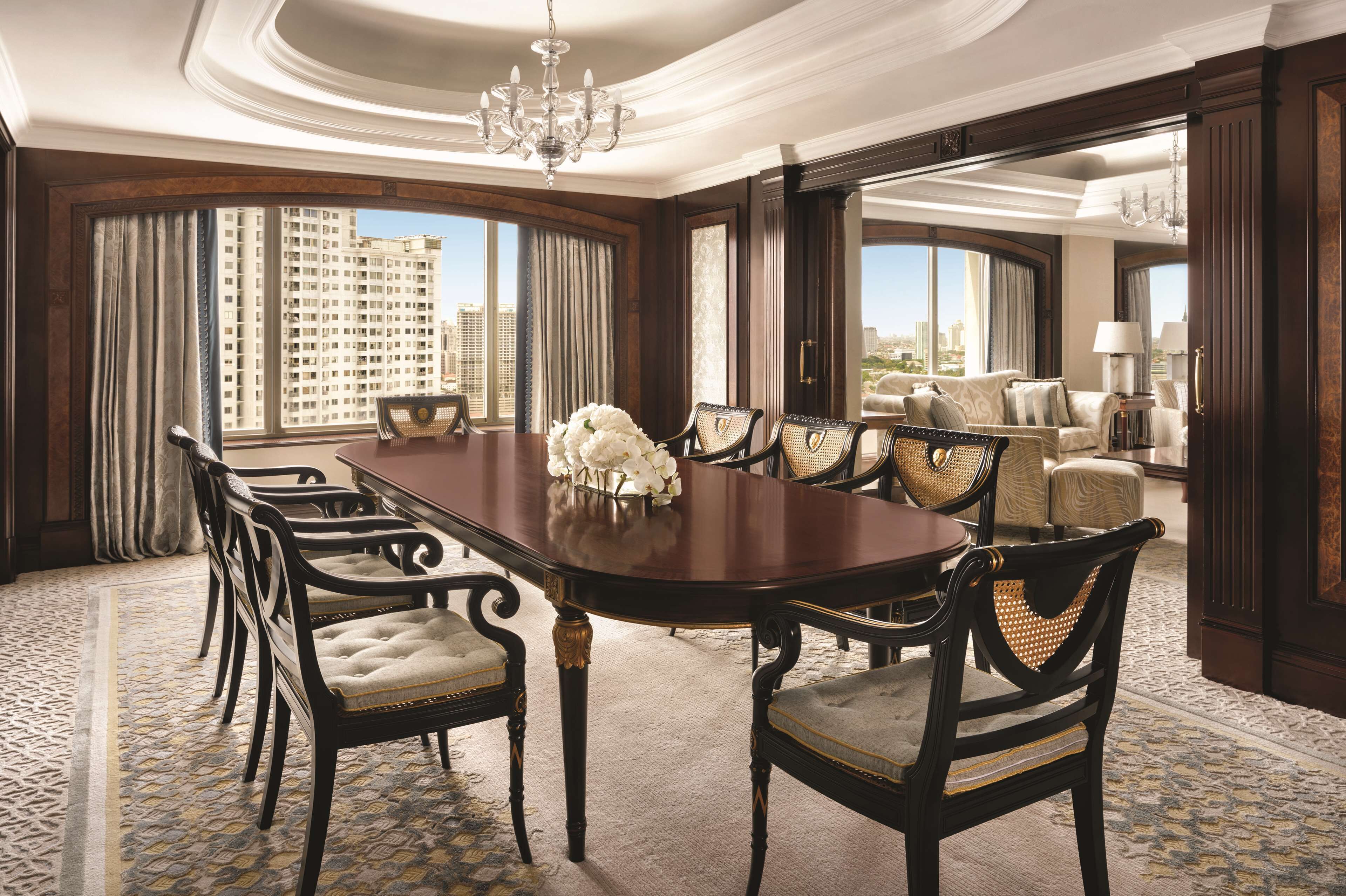 A spacious dining area with a long table and chairs, large windows with curtains, and a view of a cityscape.