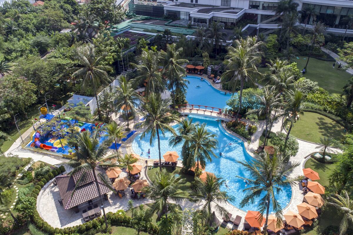 An aerial view of a resort with a central swimming pool surrounded by palm trees and sun loungers.