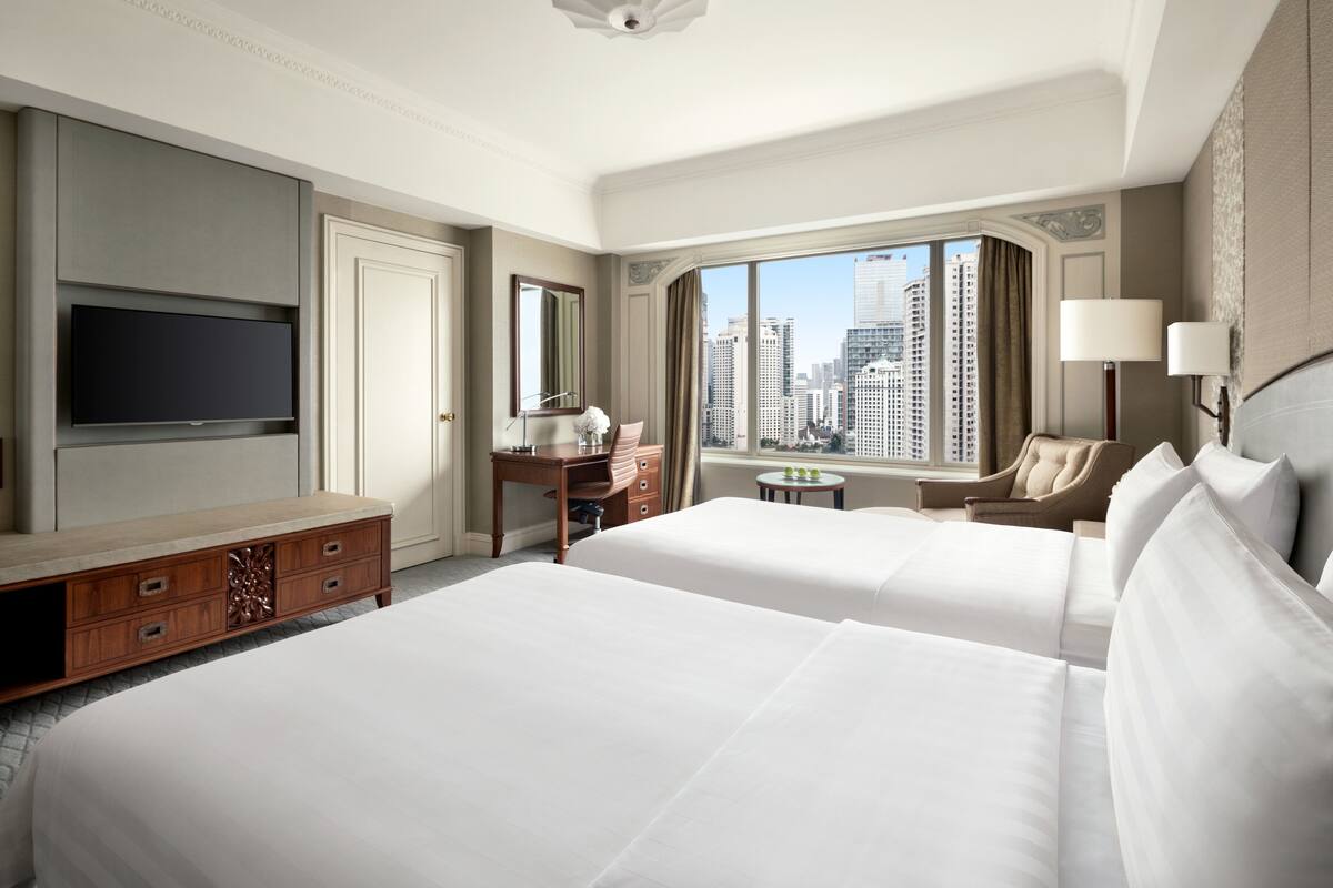 A hotel room with two beds, a TV, a desk, a chair, and a view of the cityscape.