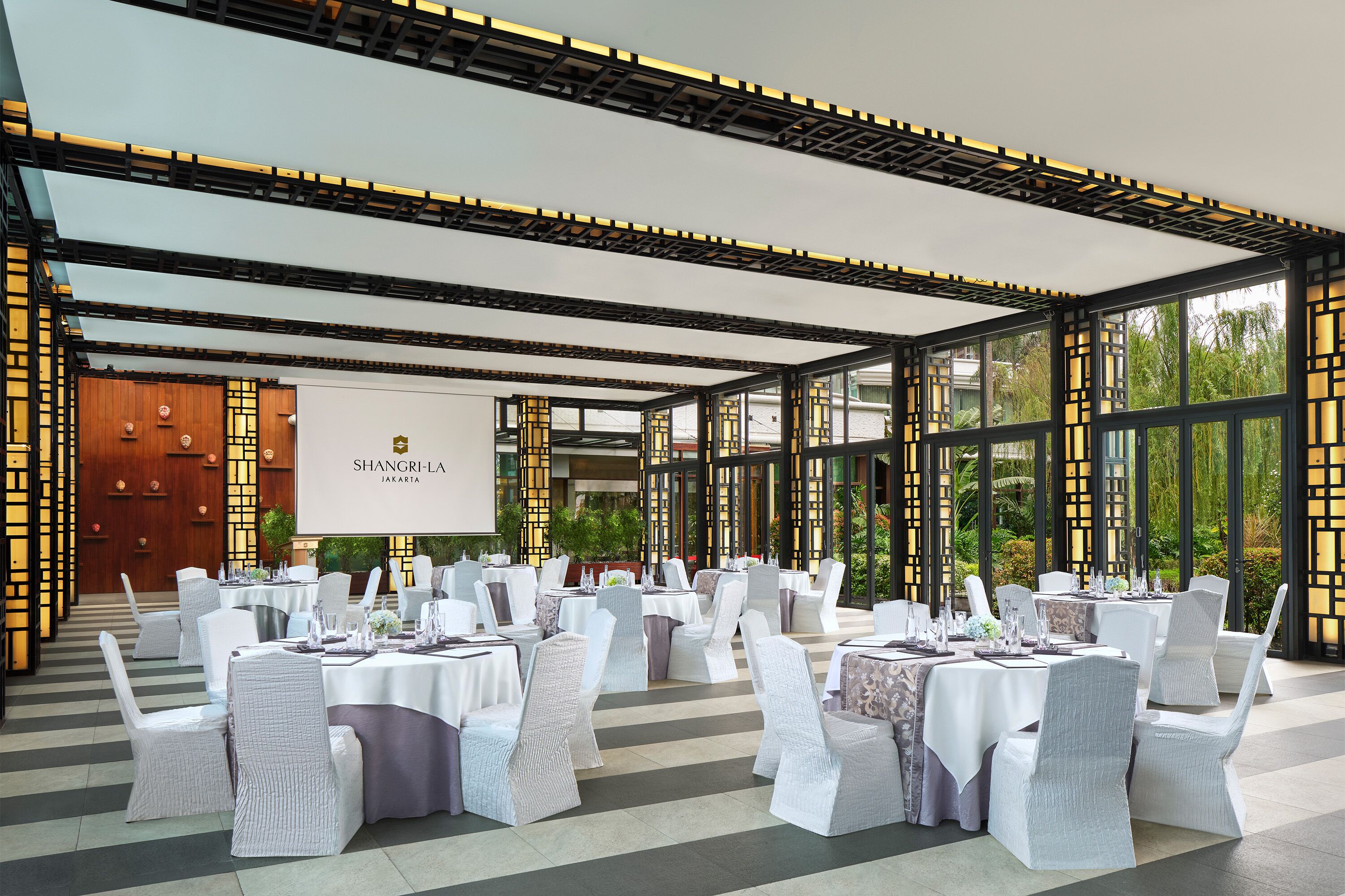 A banquet hall with round tables, white chairs, and a large screen displaying "SHANGRI-LA JAKARTA".