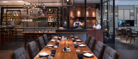 A modern restaurant with a long wooden table set for dining, a bar area with shelves of bottles, and a chef preparing food in the background.