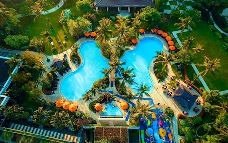 An aerial view of a resort with a large central swimming pool, surrounded by palm trees and outdoor seating areas.