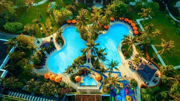 Vista aérea de un resort con una amplia piscina central, rodeado de palmeras y zonas de estar al aire libre.