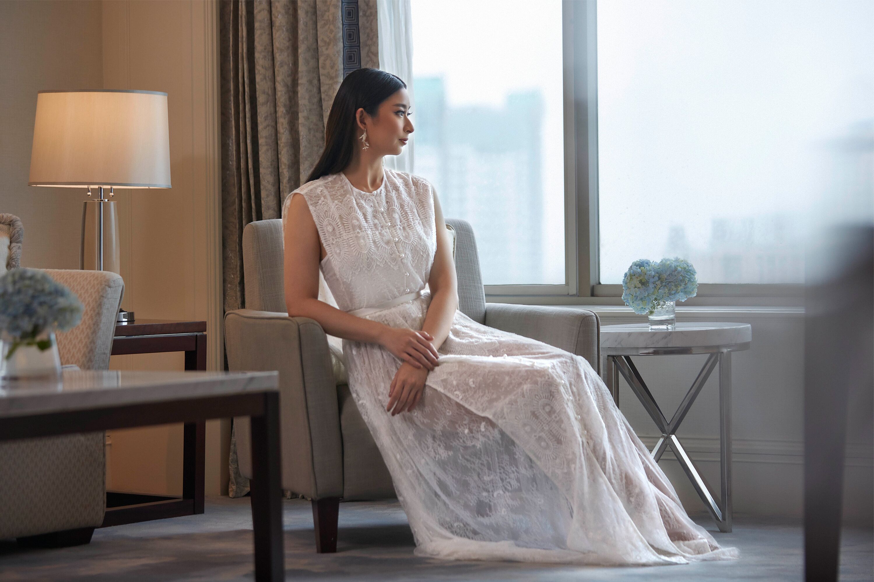 A woman in a white dress sitting by a window with a view of a cityscape.
