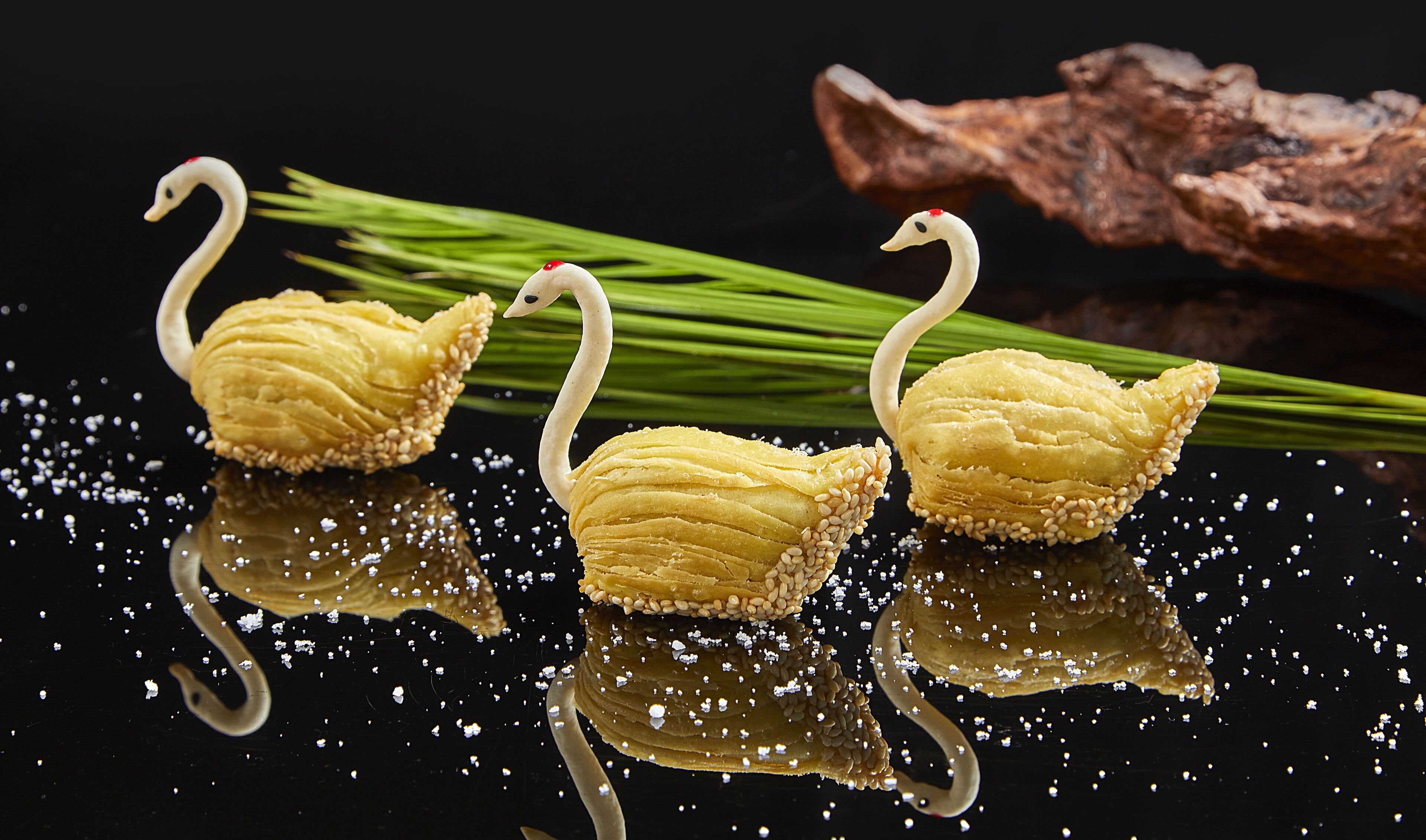 Three swan-shaped pastries on a reflective surface with green onions and a piece of wood in the background.