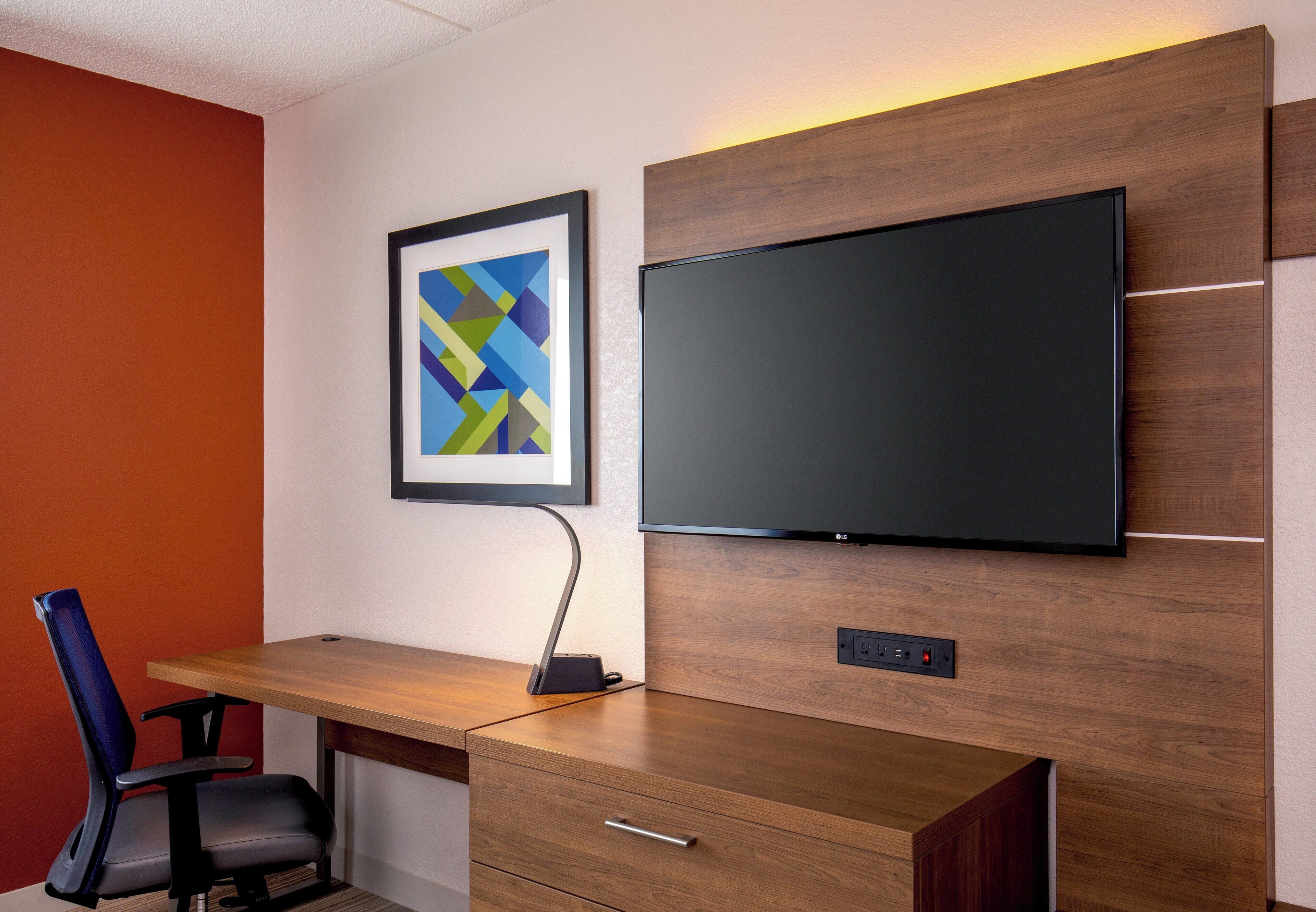 Een moderne kantoorruimte met een houten bureau, een zwarte stoel, een lijst met abstracte kunst en een wandgemonteerde flatscreen-televisie.