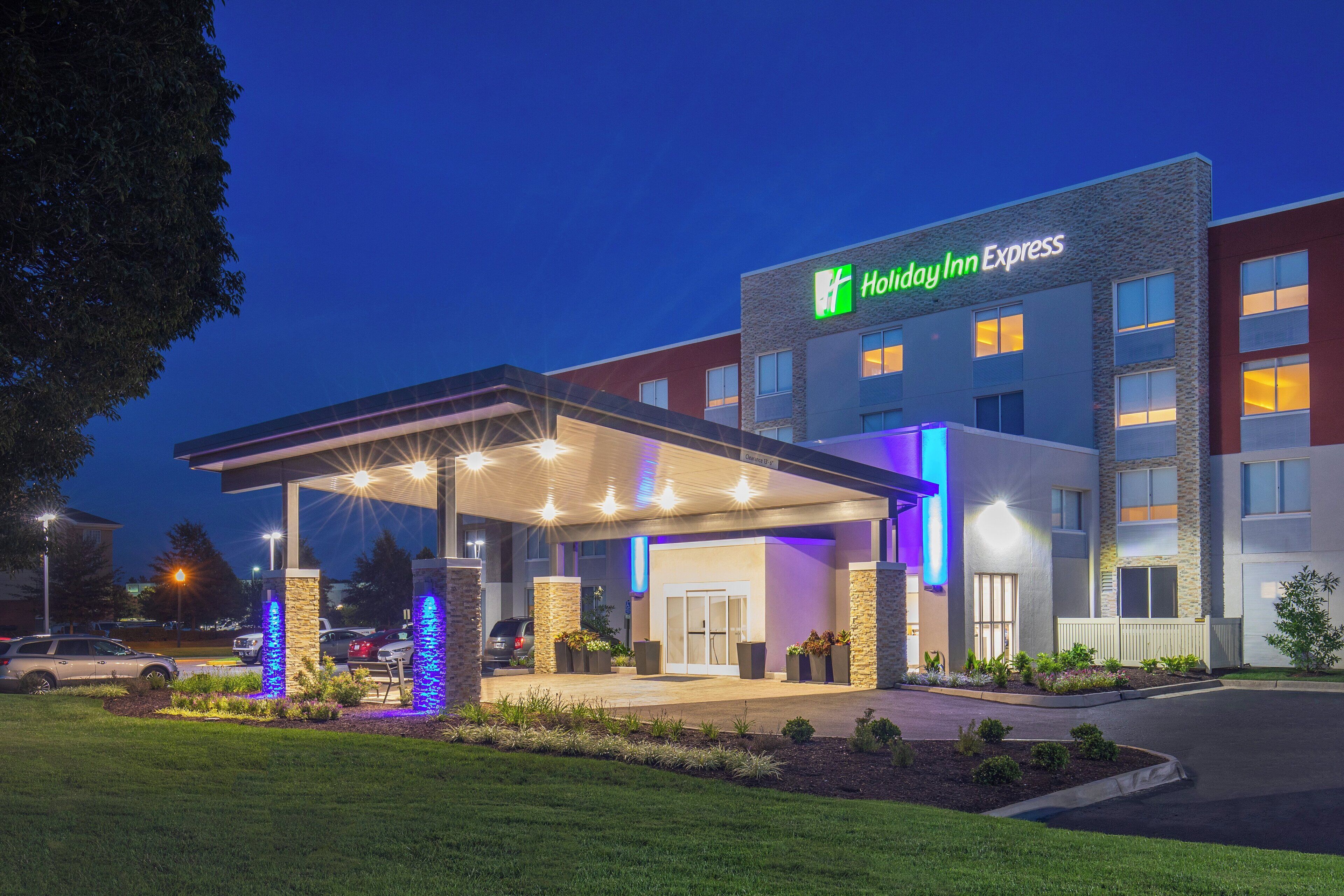 Un hotel Holiday Inn Express con entrada cubierta, iluminación exterior y una zona ajardinada con plantas.