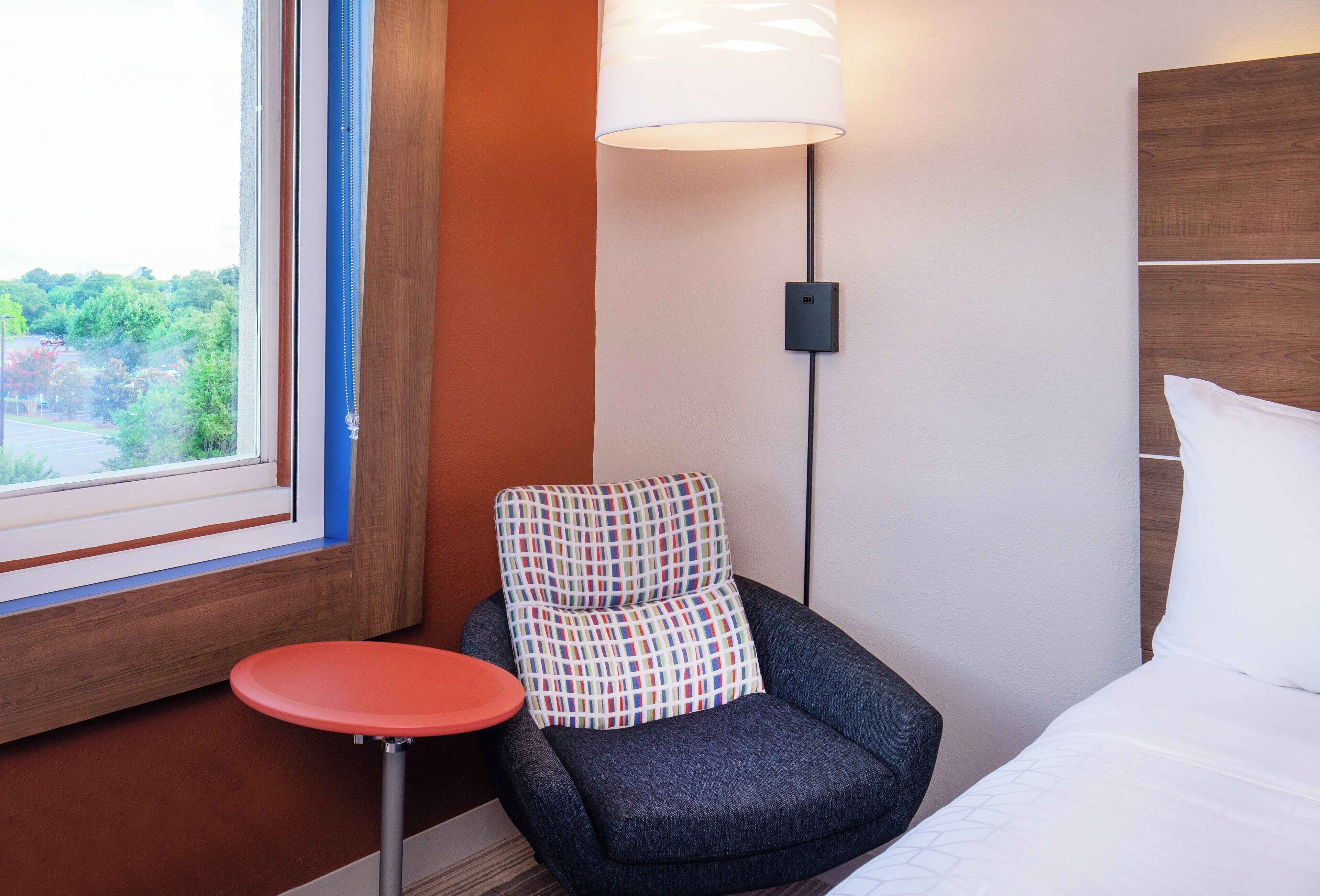 A hotel room with a window, a small round table, a chair with a colorful cushion, a wall-mounted lamp, and a bed with white bedding.