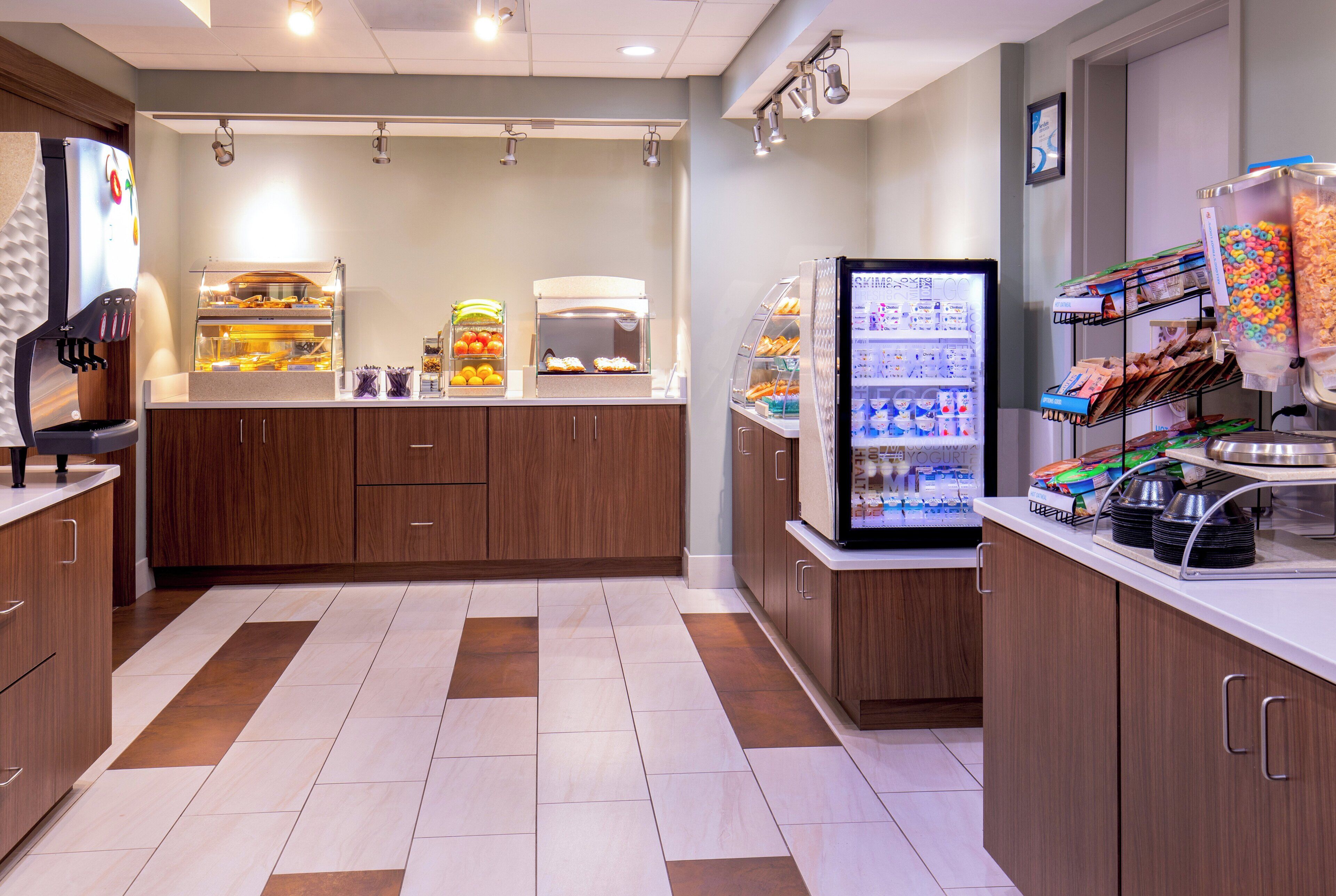 A self-service breakfast area with a coffee machine, fruit display, yogurt, cereal, and other breakfast items.