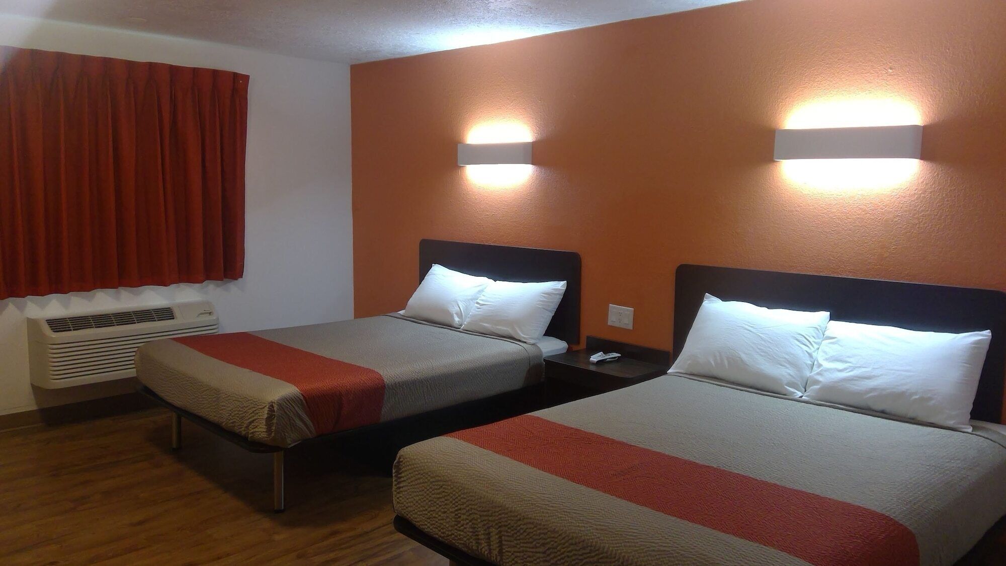 Una camera d'albergo con due letti, ognuno con testiera, una finestra con tende rosse e un condizionatore d'aria.
