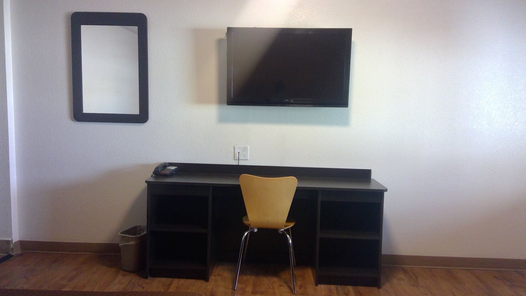 Una habitación con escritorio, silla, televisor en la pared y un cubo de basura.