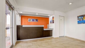 Un área de recepción con un mostrador oscuro, una pared blanca con un cartel, y suelo de baldosas.