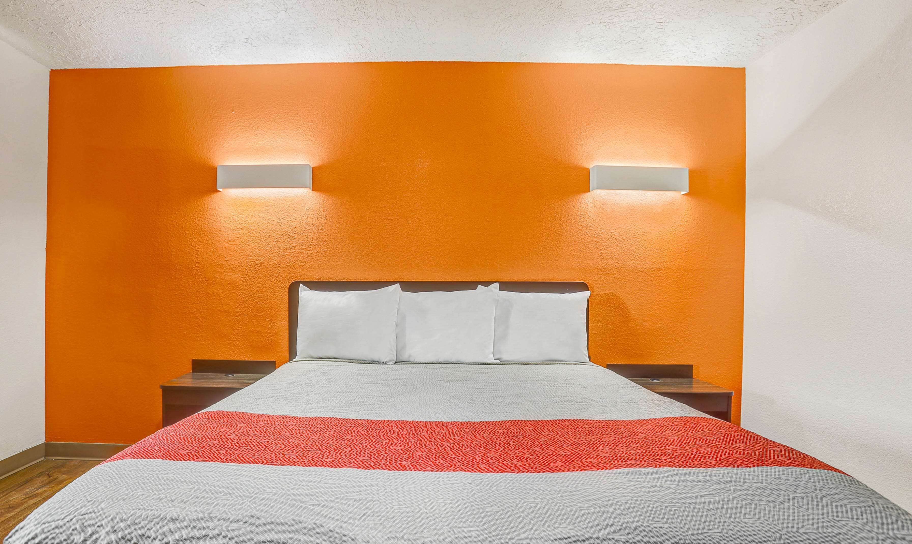 A hotel room with an orange accent wall, a bed with white and gray bedding, and two wall-mounted lights.