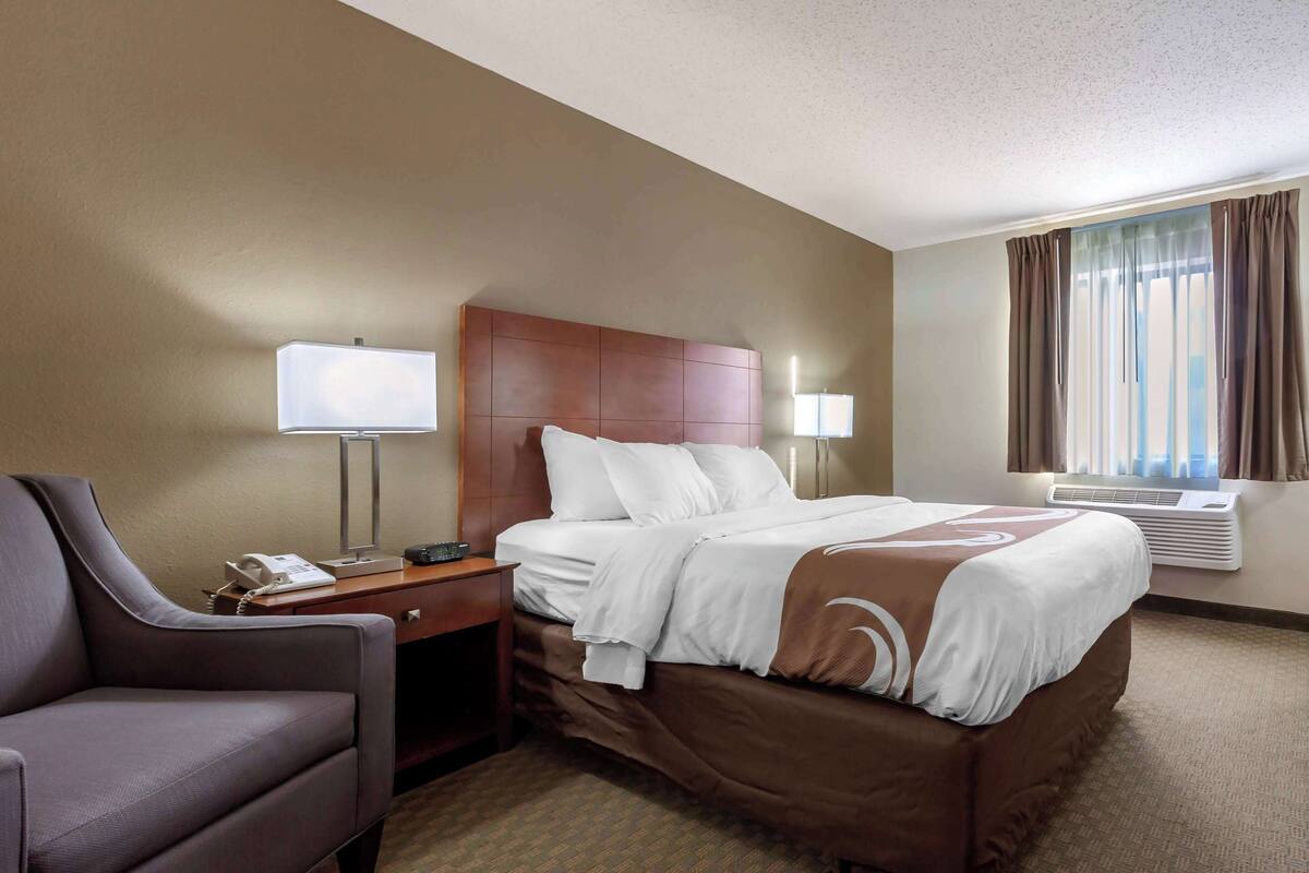 A hotel room with a bed, a desk, a chair, a lamp, and an air conditioning unit.