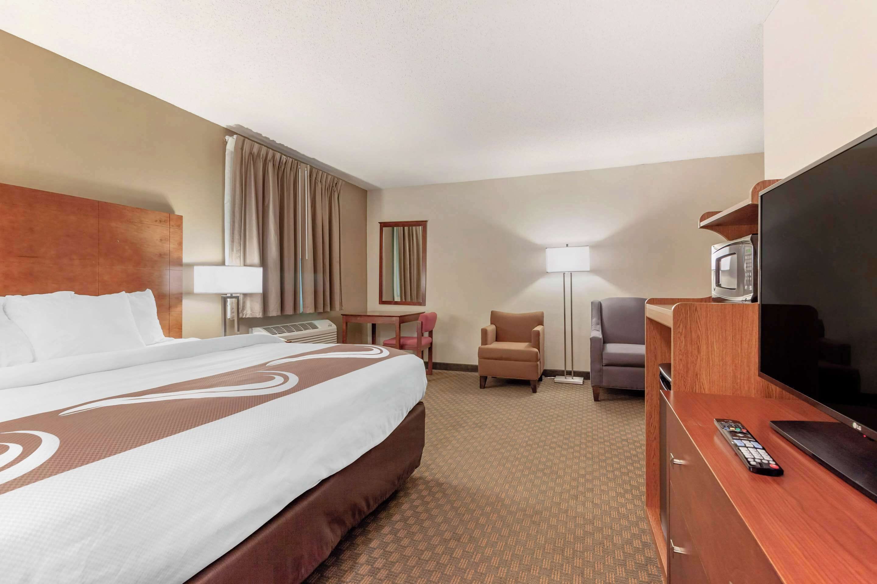 A hotel room with a large bed, a desk with a lamp, a chair, a TV, and a remote control.
