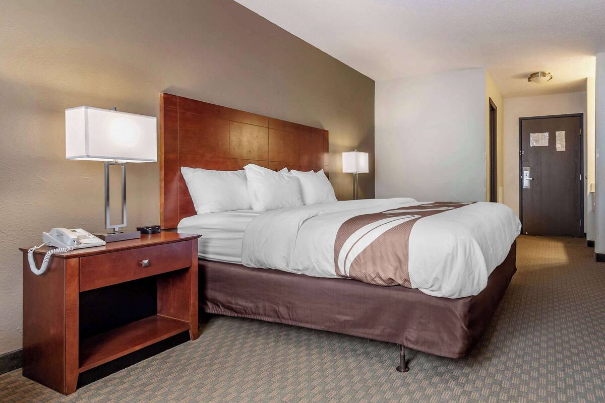 A hotel room with a large bed, a wooden nightstand with a phone, two bedside lamps, and a carpeted floor.