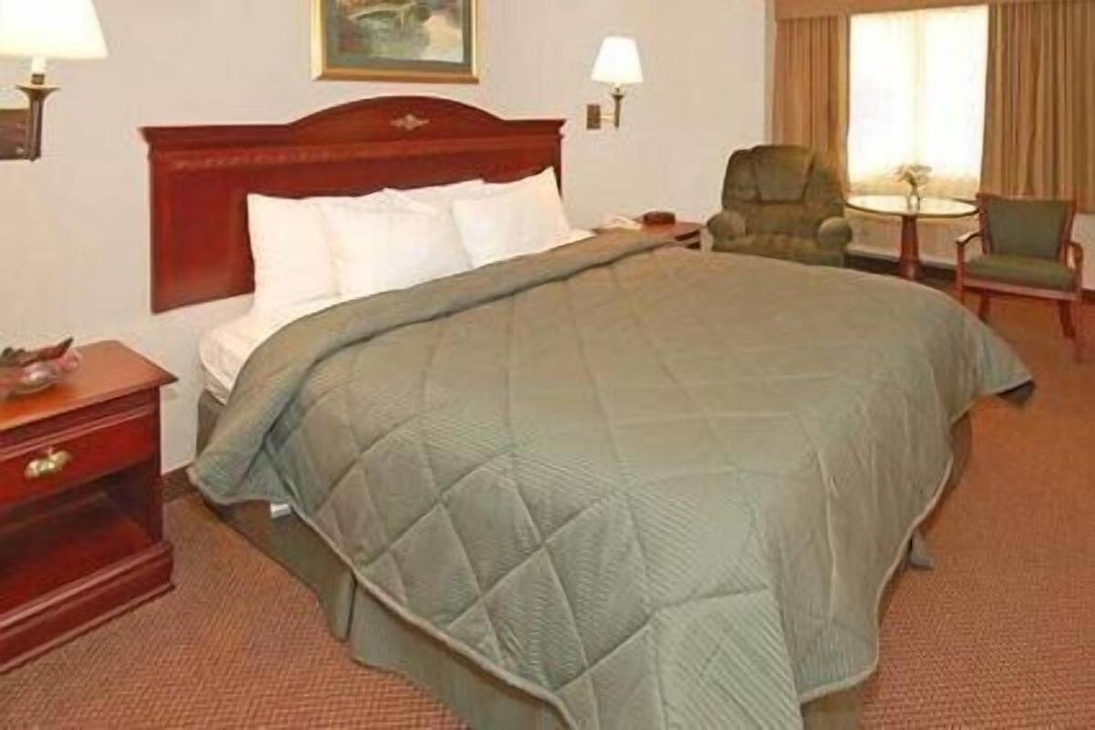 Quarto de hotel com uma cama grande, duas mesas de cabeceira, uma cadeira, uma mesinha e um quadro na parede.